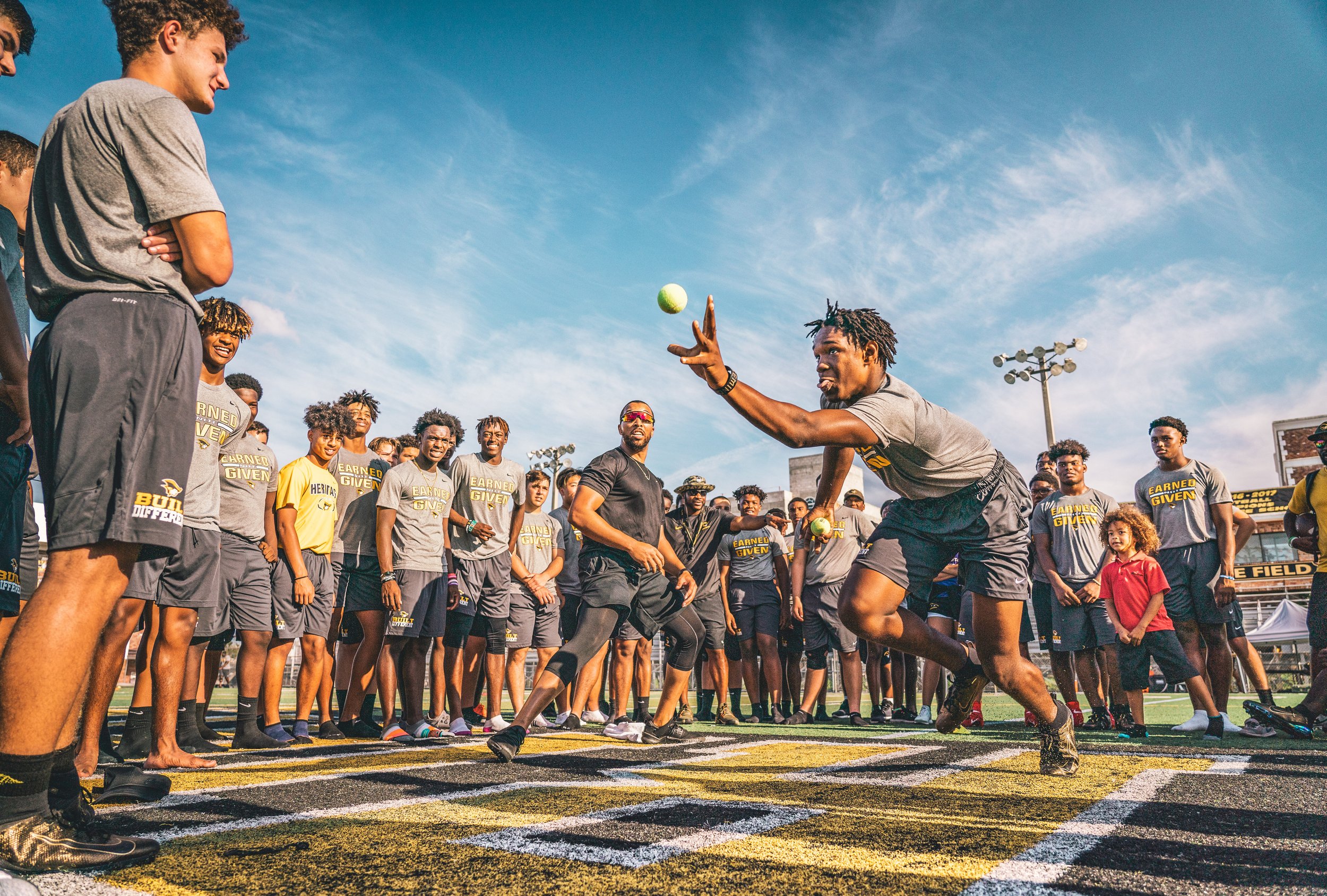 Group of male athletes in sportswear participating in an outdoor training activity, one person reaching out to catch a ball, surrounded by others observing on a sports field.