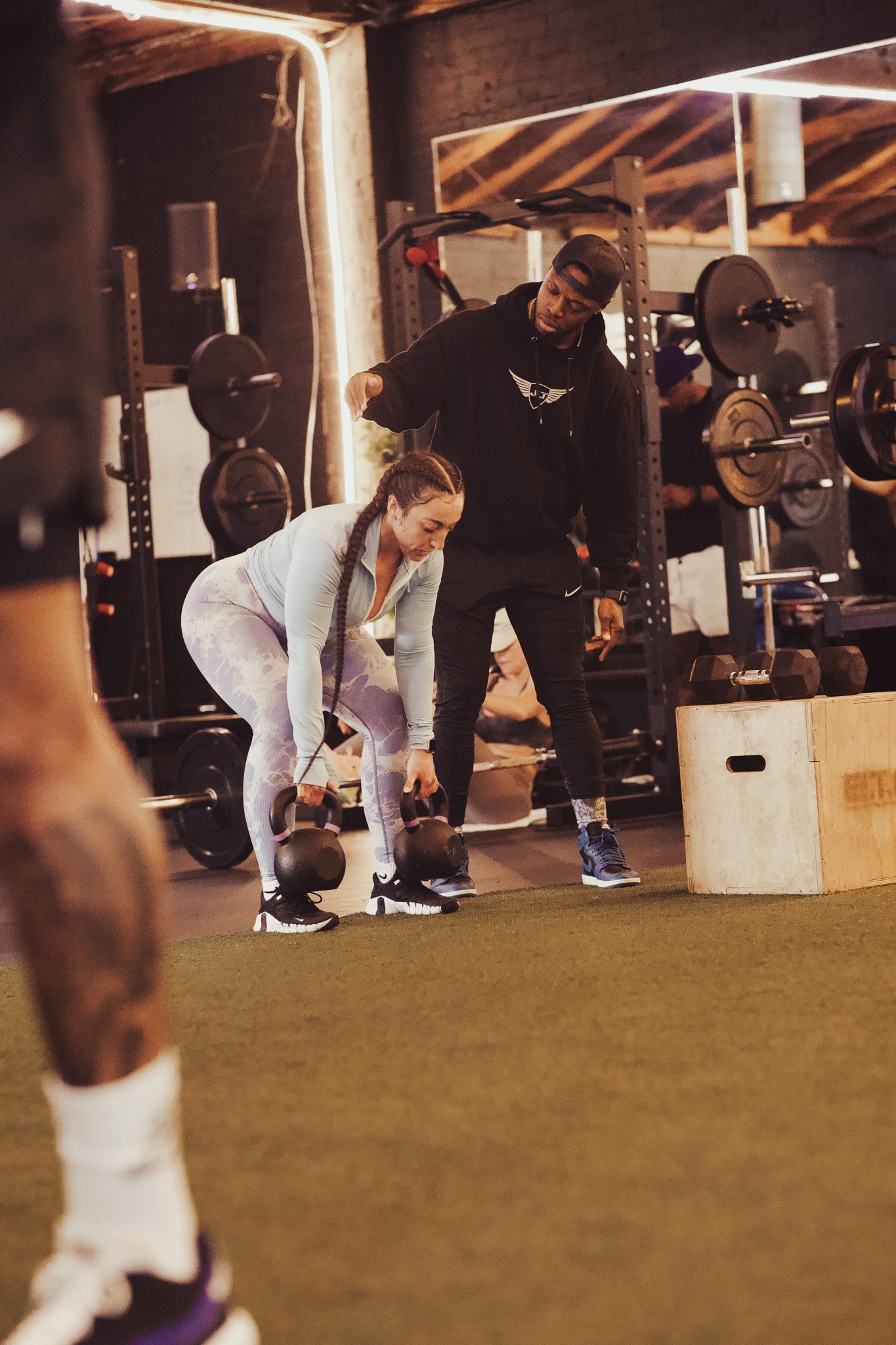 A woman performing a kettlebell deadlift in a gym under the guidance of a trainer.