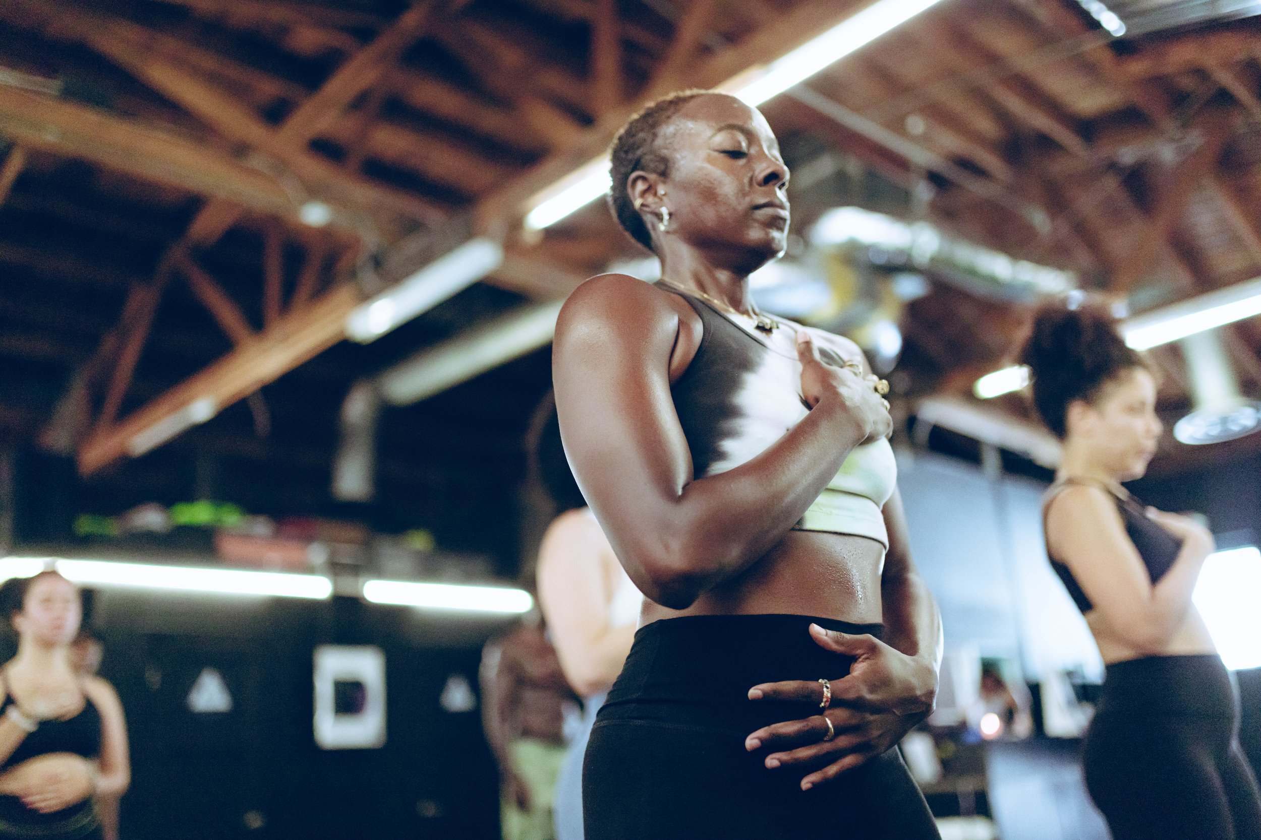 A group of women practicing yoga indoors, focusing on mindfulness and breathwork. The setting appears to have industrial-style lighting and exposed beams. The women are wearing activewear, with their hands placed on their chests and stomachs.