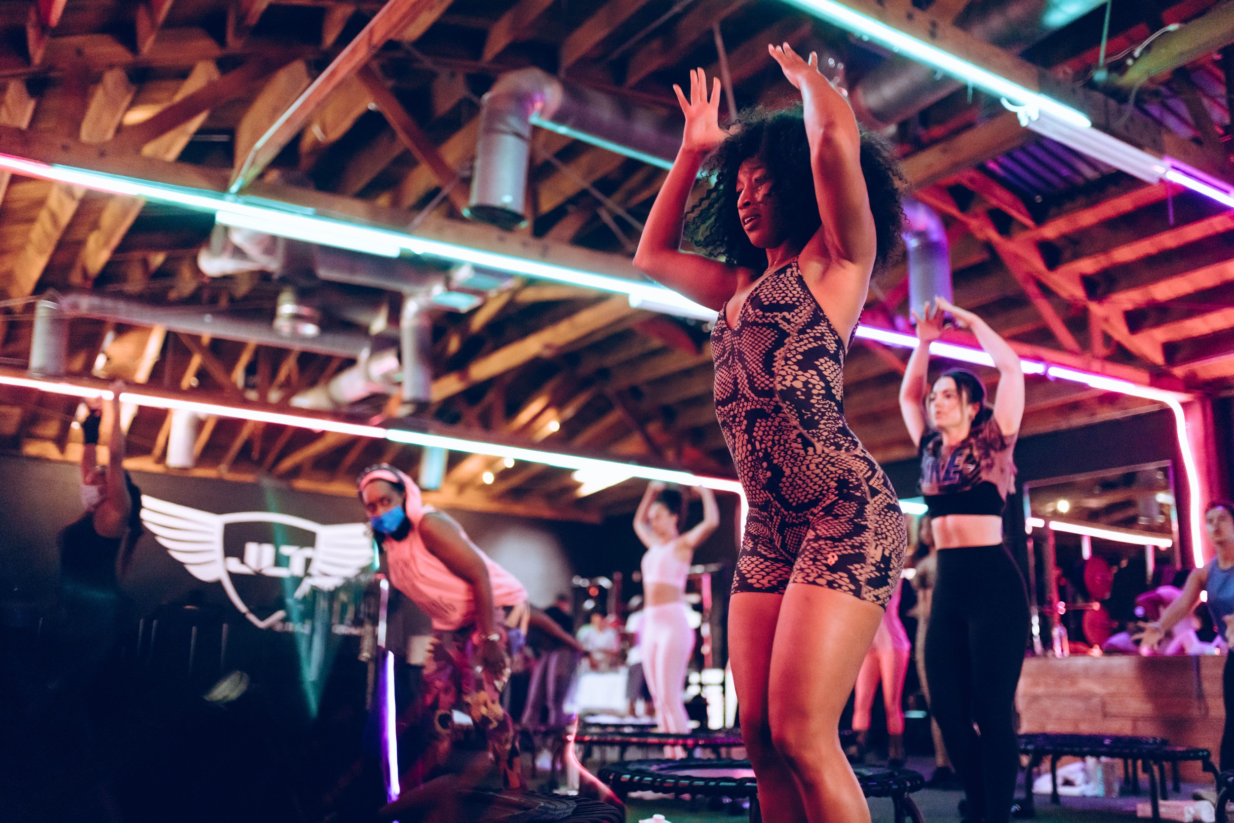 A group of people participating in a dance fitness class, with colorful neon lights and wooden beams in the ceiling.