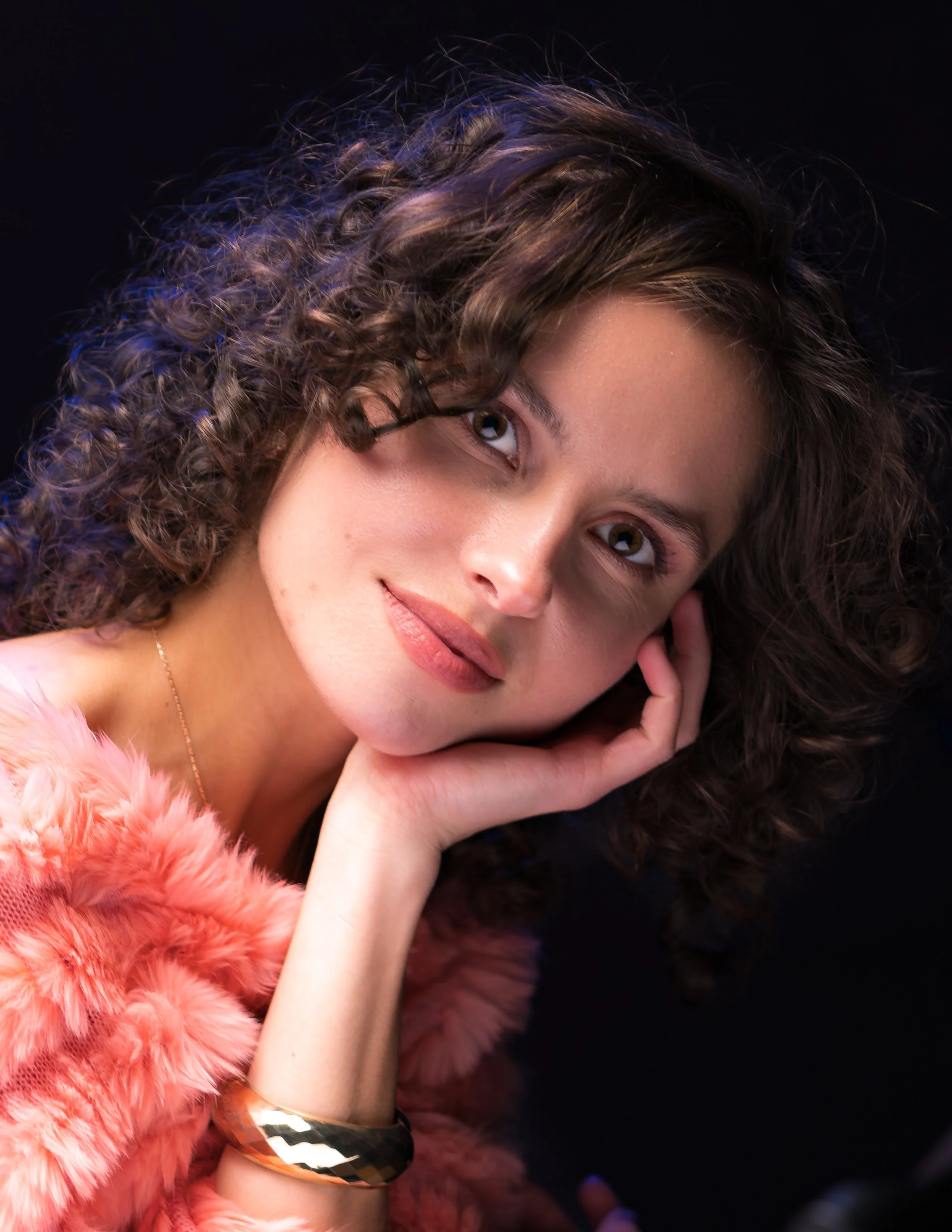 A close-up portrait of a woman with curly brown hair, smiling softly, wearing a pink faux fur garment and a reflective bracelet, against a dark background.