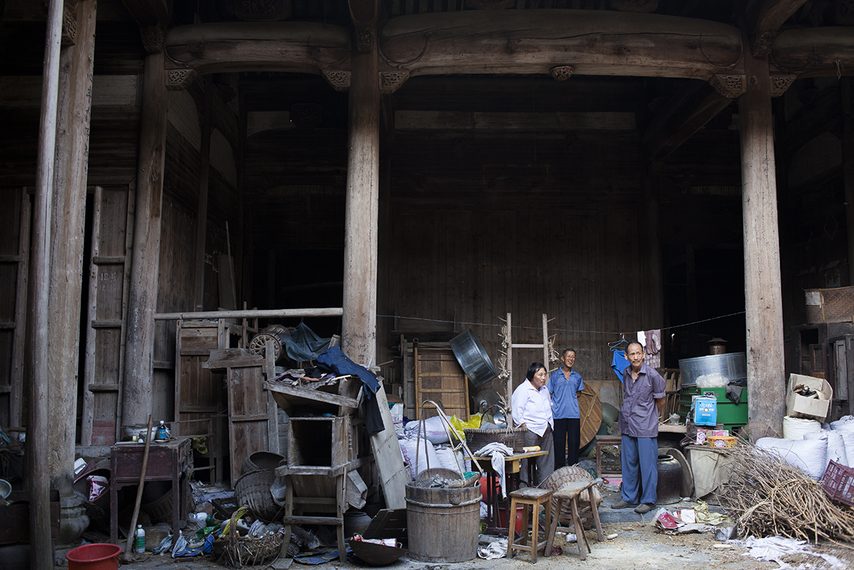   Wang Shouchang (right) looks out at the main courtyard of Mingxian Hall with its caretaker, Wang Changfa (center), 72, and his wife. Above the clutter, the elegant timbered roof beams remain intact. Wang Changfa began renting Mingxian hall from the