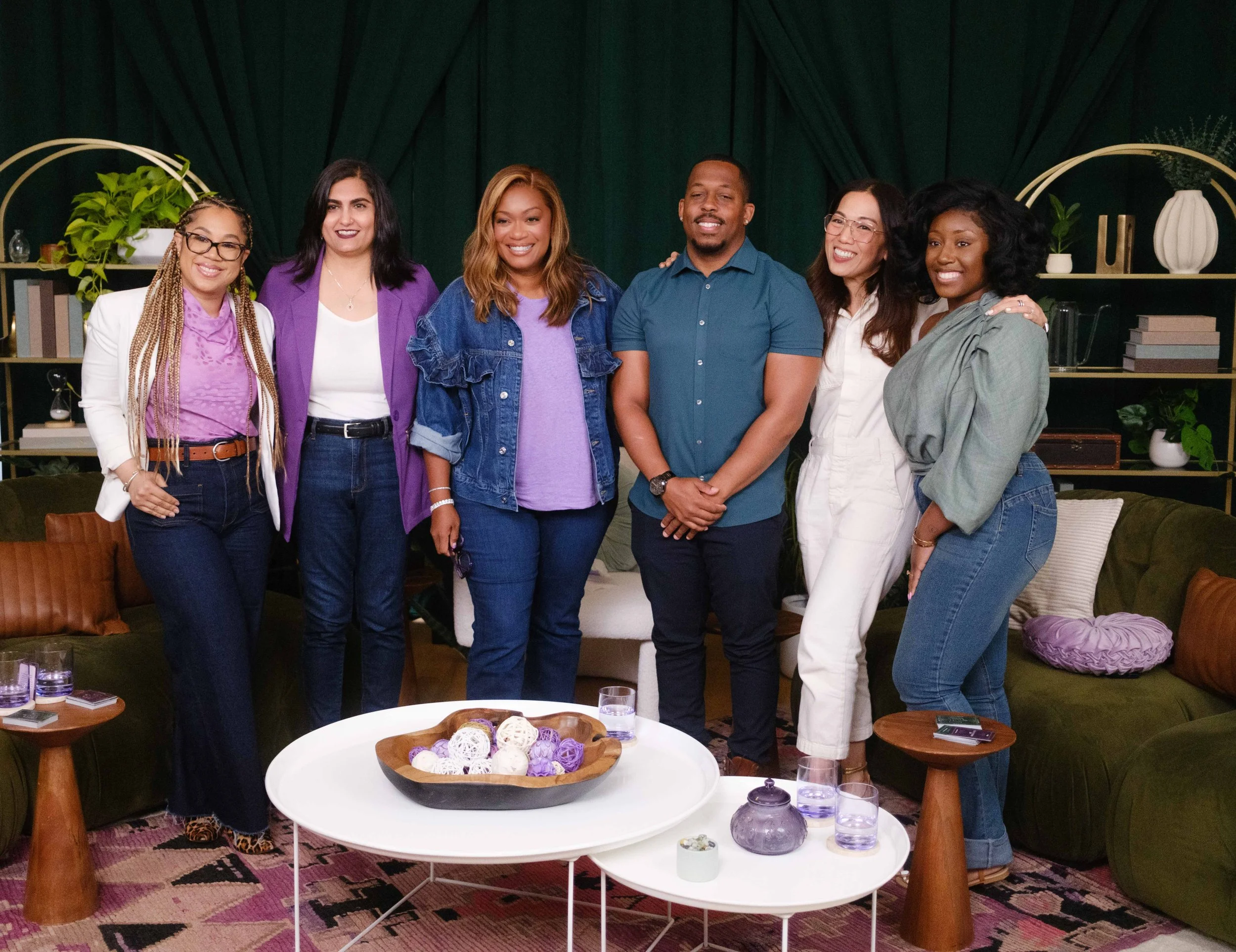 Group of seven diverse people smiling and posing together indoors, with dark green curtains and decorative shelves with plants and books in the background.