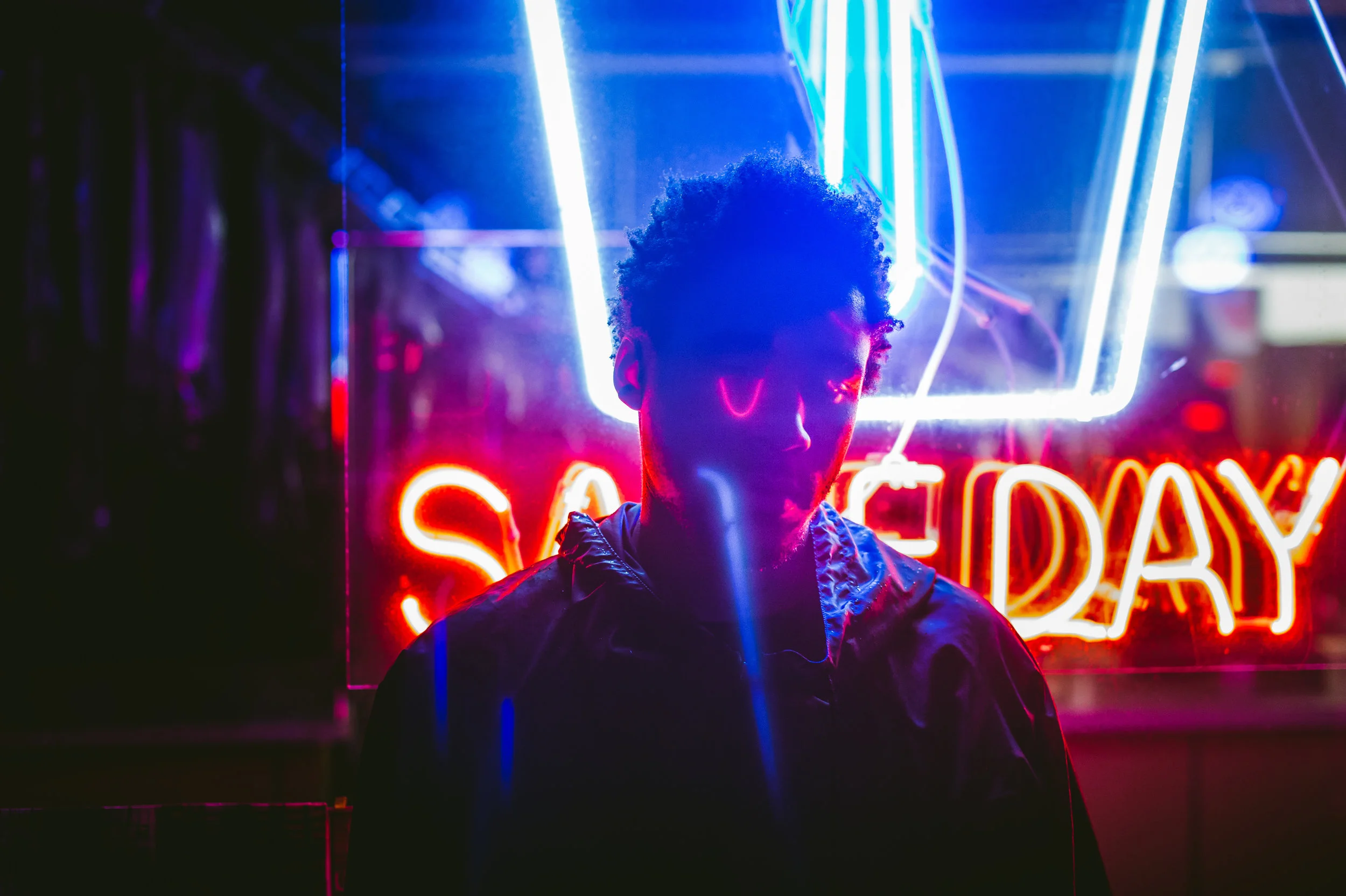 A person stands in front of a neon sign that reads 'Saturday' at night. The neon lights cast colorful glow on the person's face and jacket.