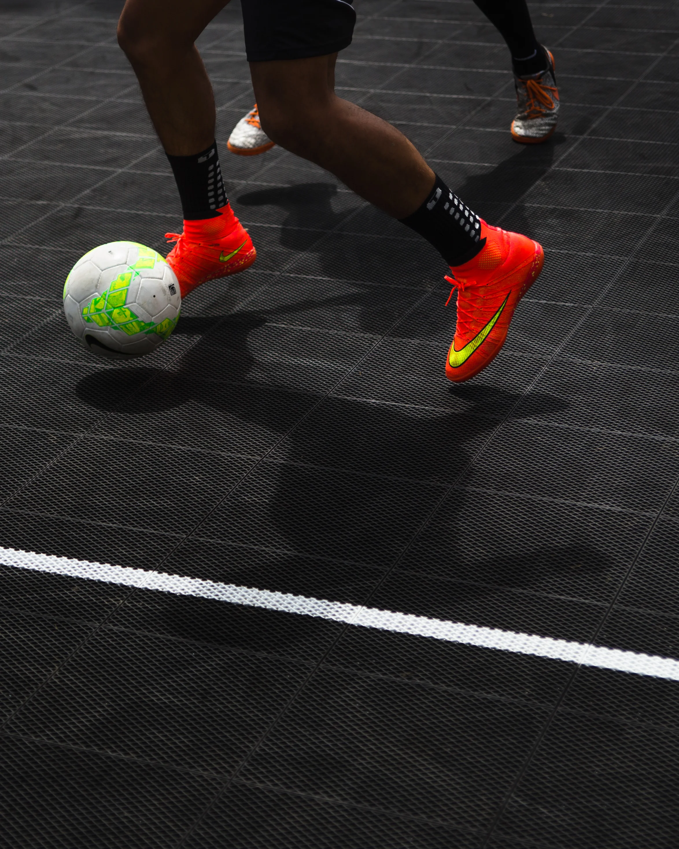Person wearing bright orange Nike soccer shoes and black socks is kicking a white and green soccer ball on a black indoor court.