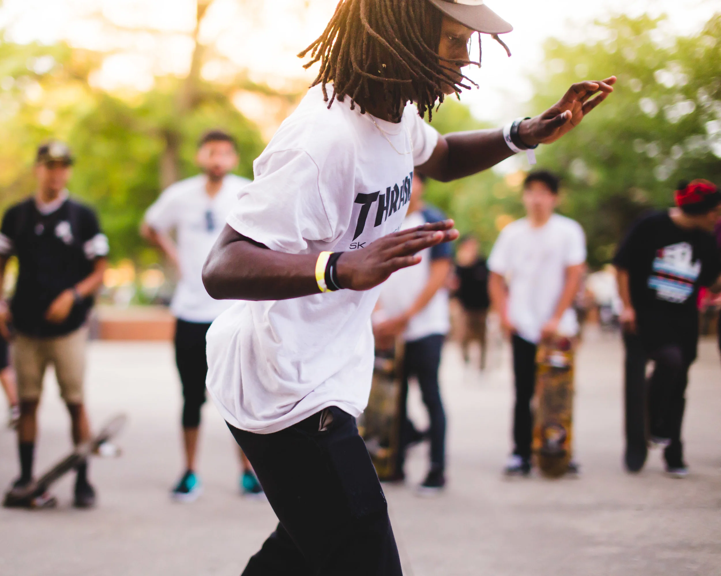 A young man with long dreadlocks and a cap dancing outdoors during a gathering, with other people watching and skateboards on the ground.
