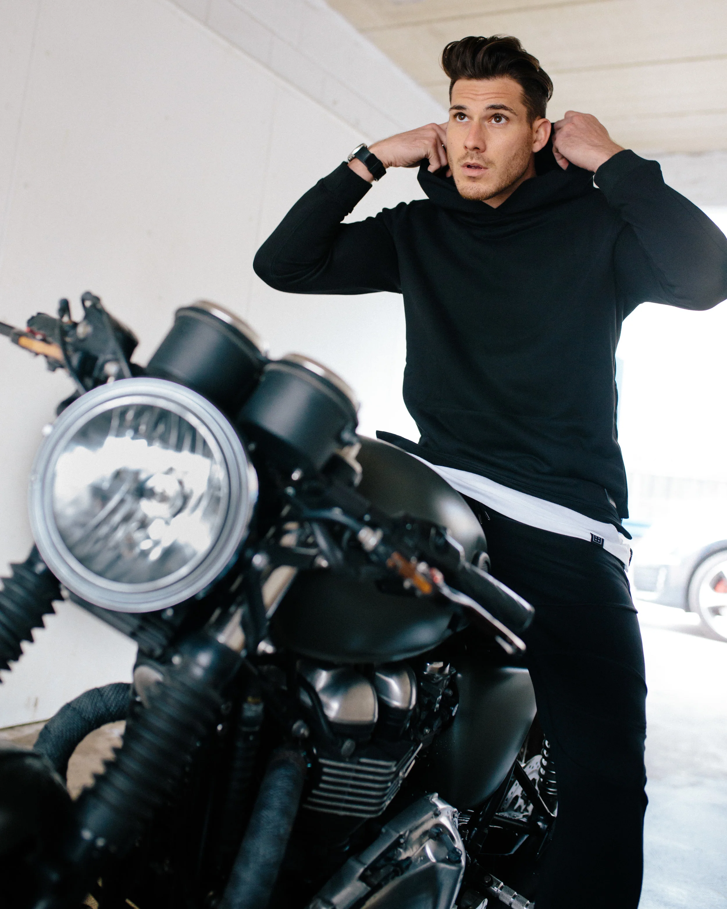 A man in a black hoodie adjusting his hood while standing next to a black motorcycle in a garage.