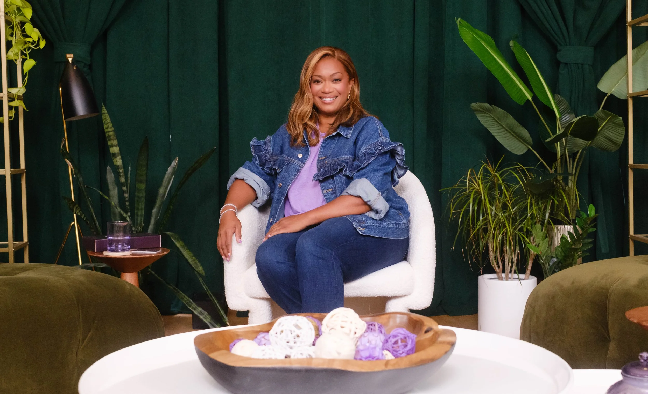 A woman with light brown hair smiling, sitting on a white armchair in a room with green curtains and several potted plants. There is a table with decorative yarn balls in front of her.