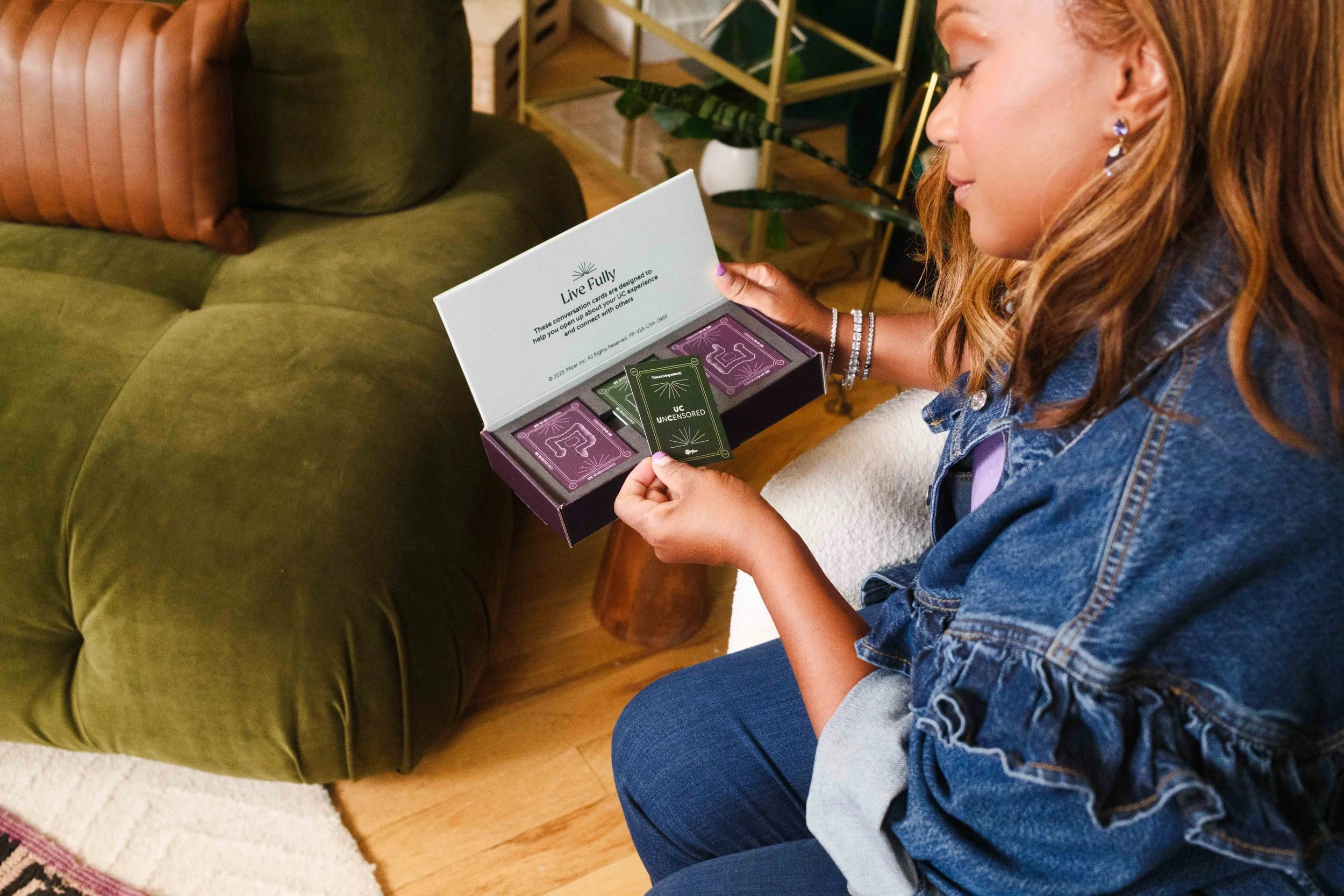 Woman sitting on a white chair, looking at a deck of cards labeled 'Live Fully' with the words 'UC UNCENSORED' on one card, and a green card in her hand, in a cozy living room with a green sofa and a gold metal plant stand in the background.