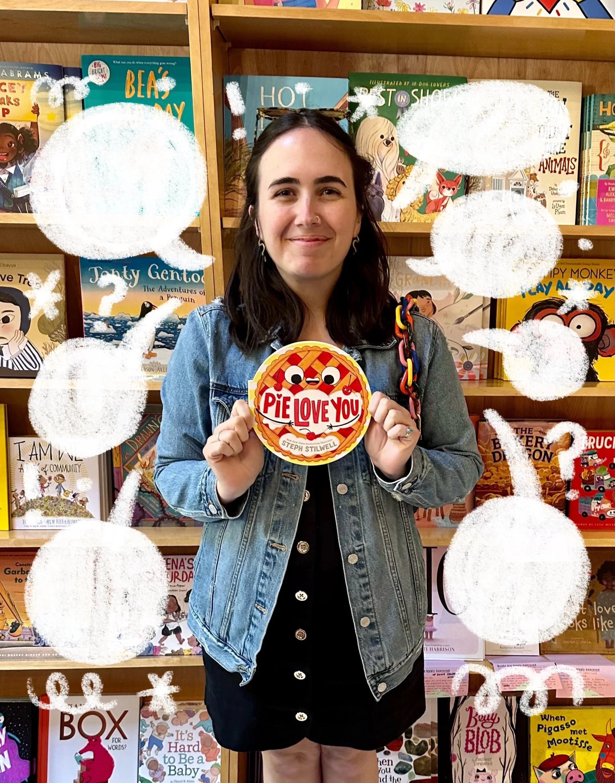 A young woman with dark hair and a denim jacket standing in front of a bookshelf filled with children's books, holding a circular book called 'Pie Love You' with a pie and two animated eyes on it.