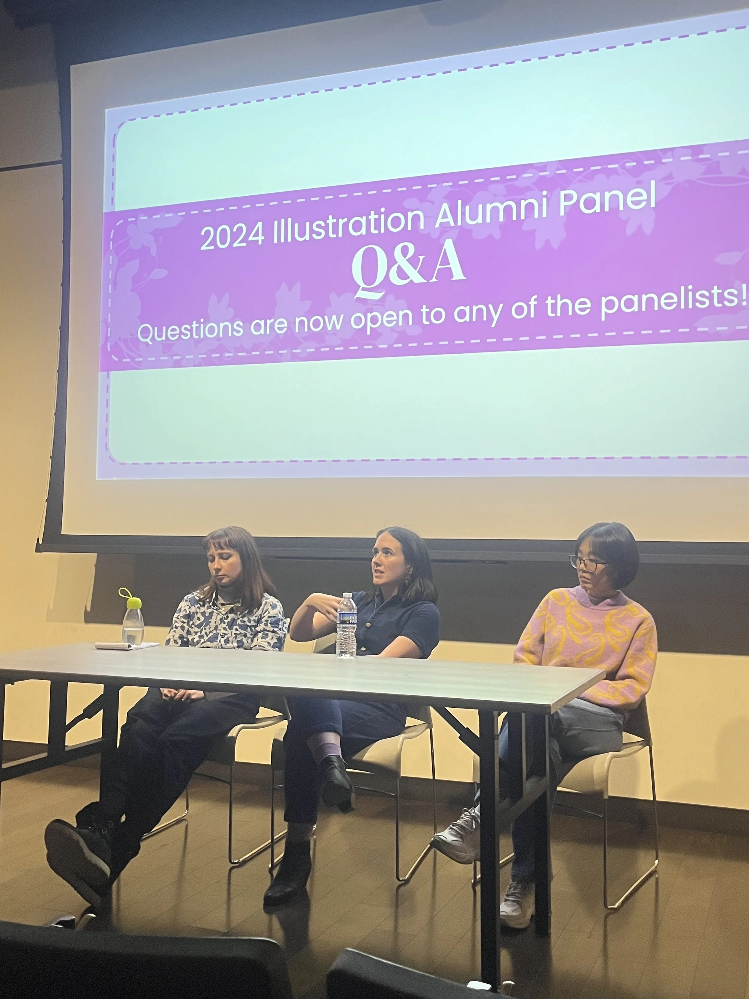 Three women sitting at a panel table during a Q&A session at a MICA Illustration Alumni Panel, with a large screen behind them displaying the event information.