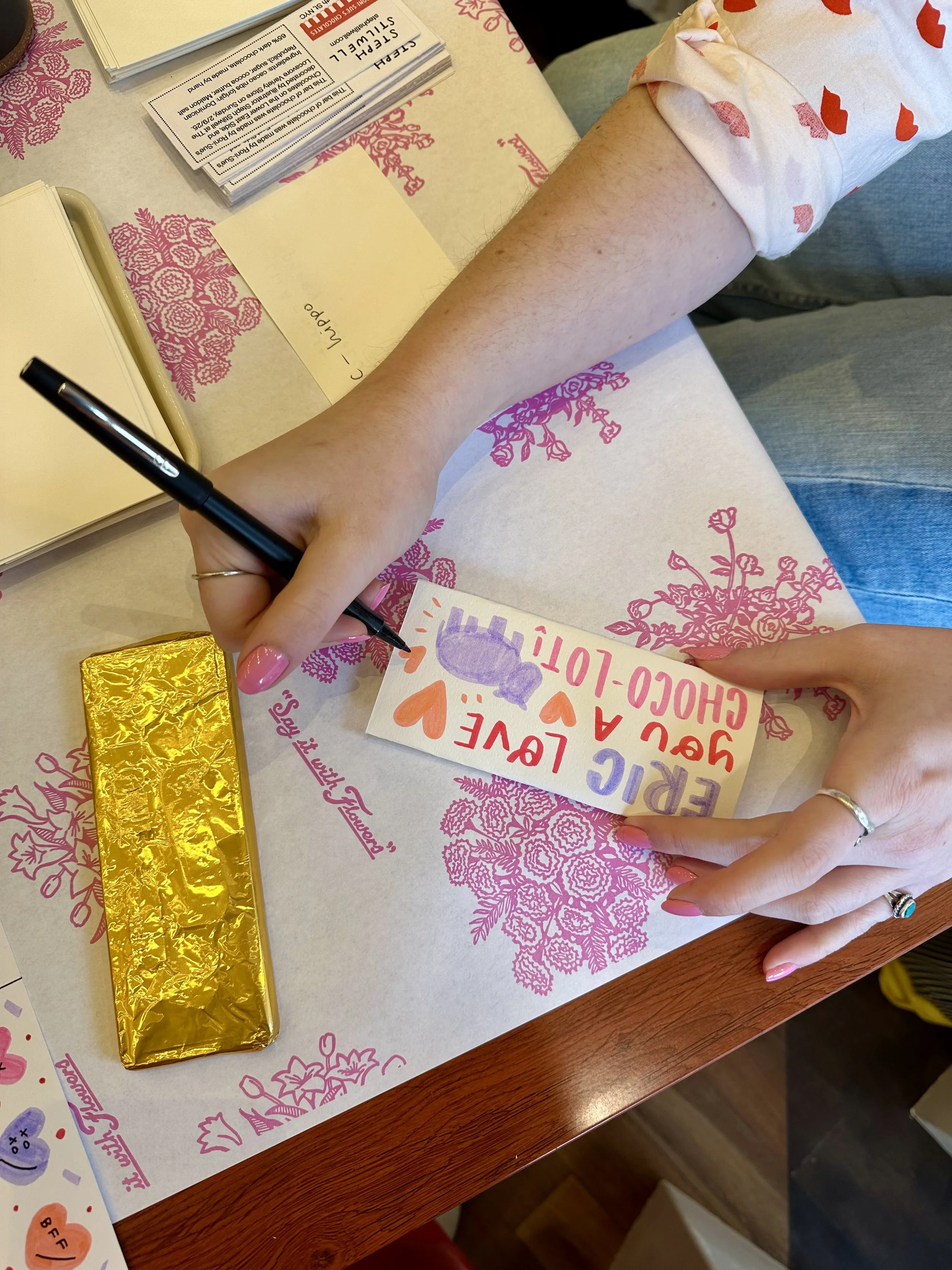 A person is customizing a candy bar wrapper with colorful hearts and text, sitting at a table with pink floral-themed paper, Valentine's Day goodies, and a gold-wrapped chocolate bar.
