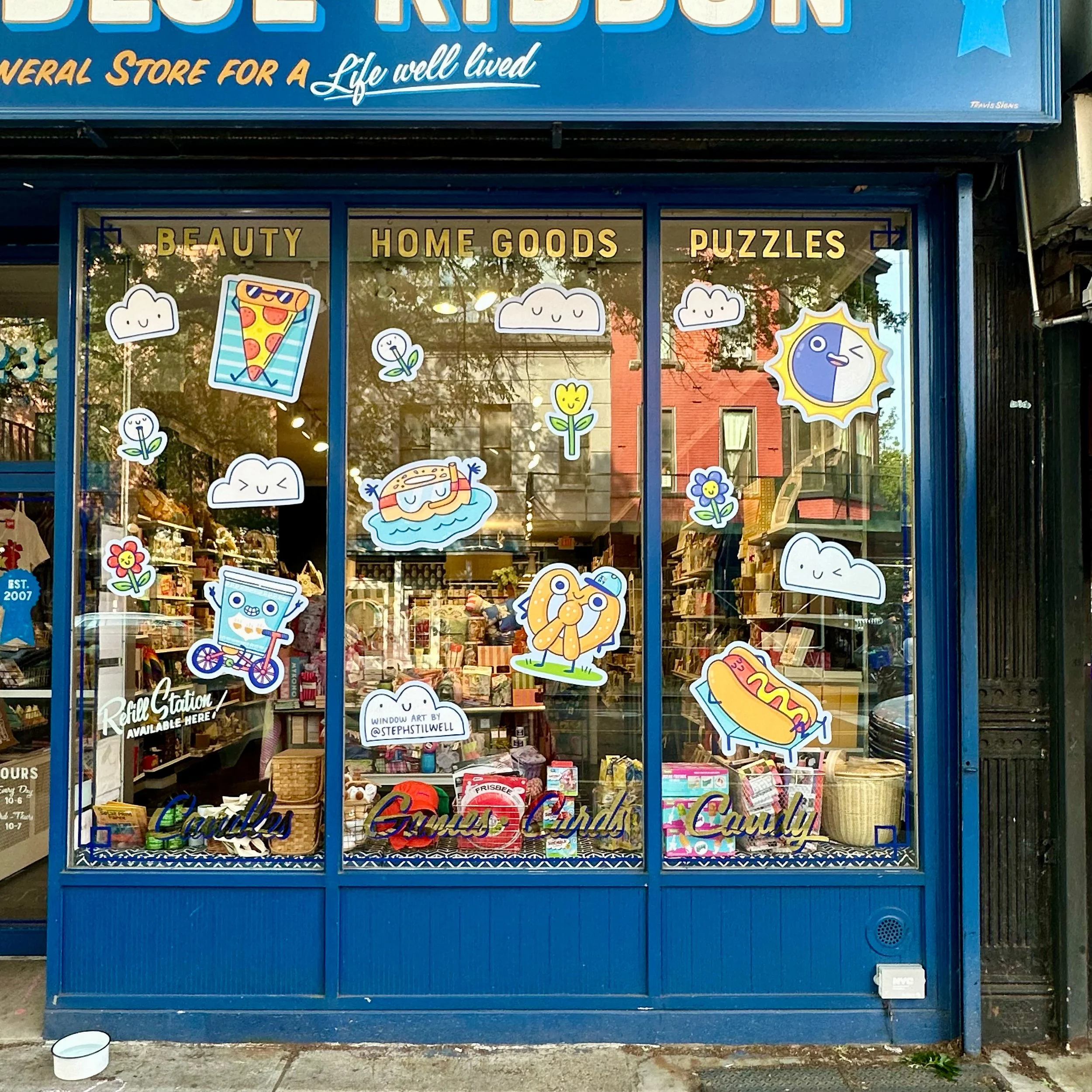 Storefront window with colorful cartoon illustrations of clouds, flowers, a pizza slice, a hot dog, a hot tub, a bicycle, and a sun, with signs reading 'Beauty', 'Home Goods', and 'Puzzles'.