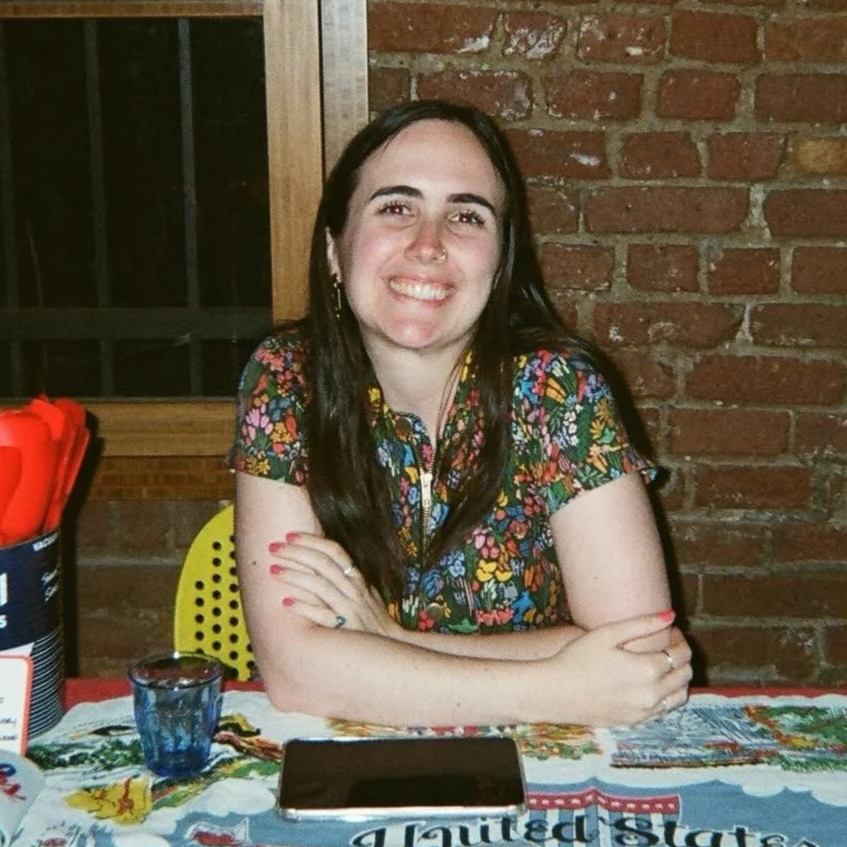 A young woman with long dark hair, smiling and sitting at a table with a colorful tablecloth. She is wearing a floral jumpsuit, with arms crossed on the table, in front of a brick wall background.