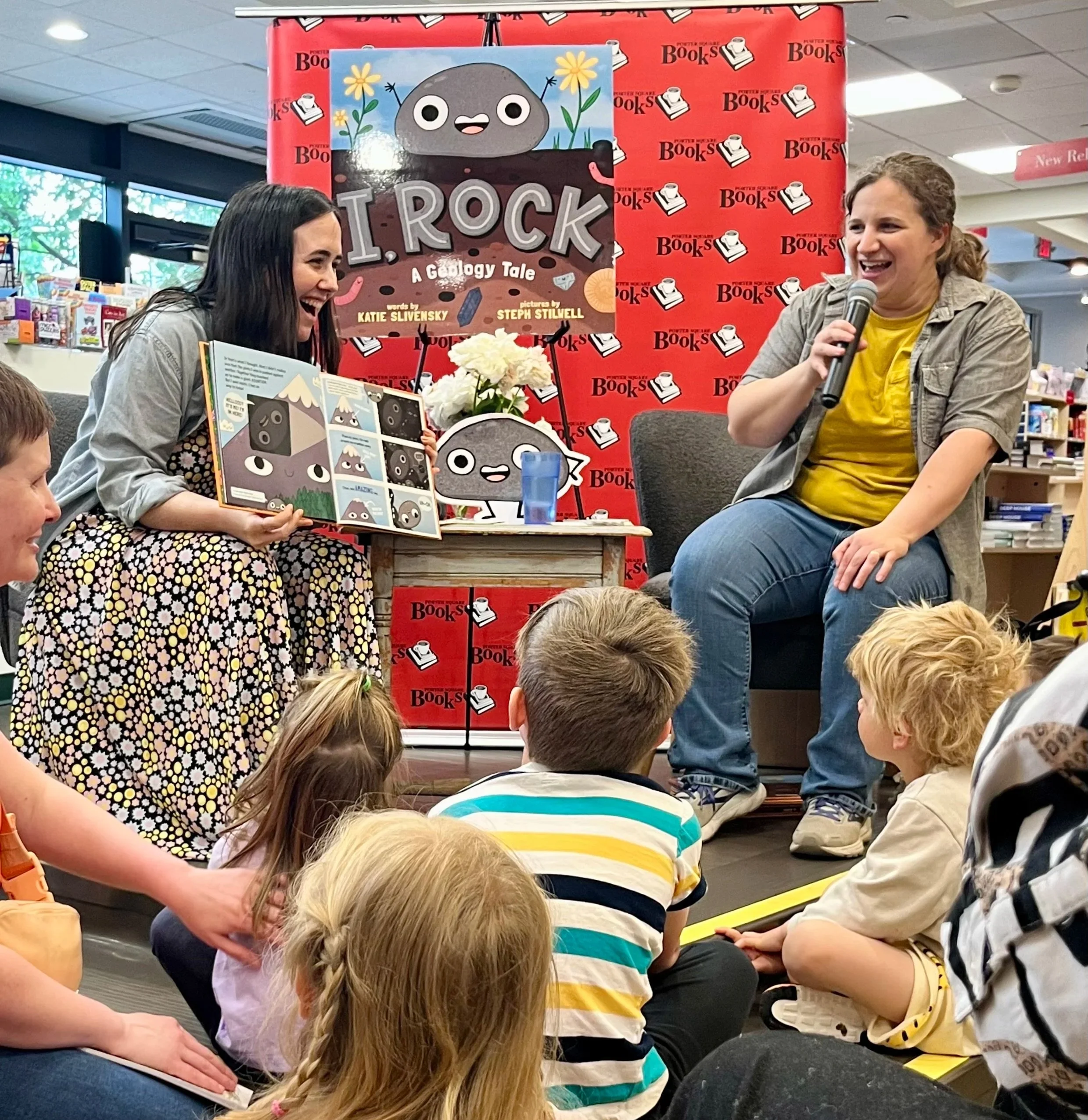 Steph is reading a children's book aloud to an audience of children at a bookstore event for a book titled 'I Rock.' The children are sitting on the floor, attentively listening, while another woman holds a microphone and interacts with the audience.