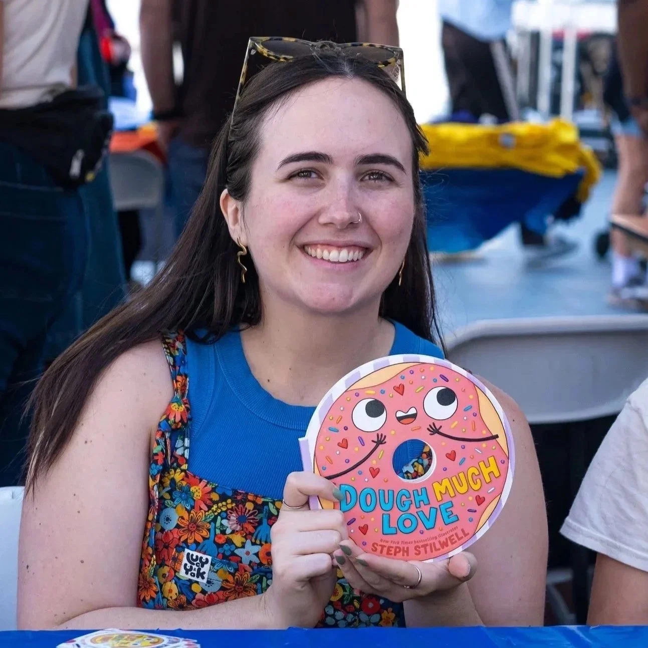 Steph, a young woman with long dark hair, smiling, holding a colorful children's book titled 'Dough Love Much' by Steph Stilwell, in a crowded setting with people and tables in the background.