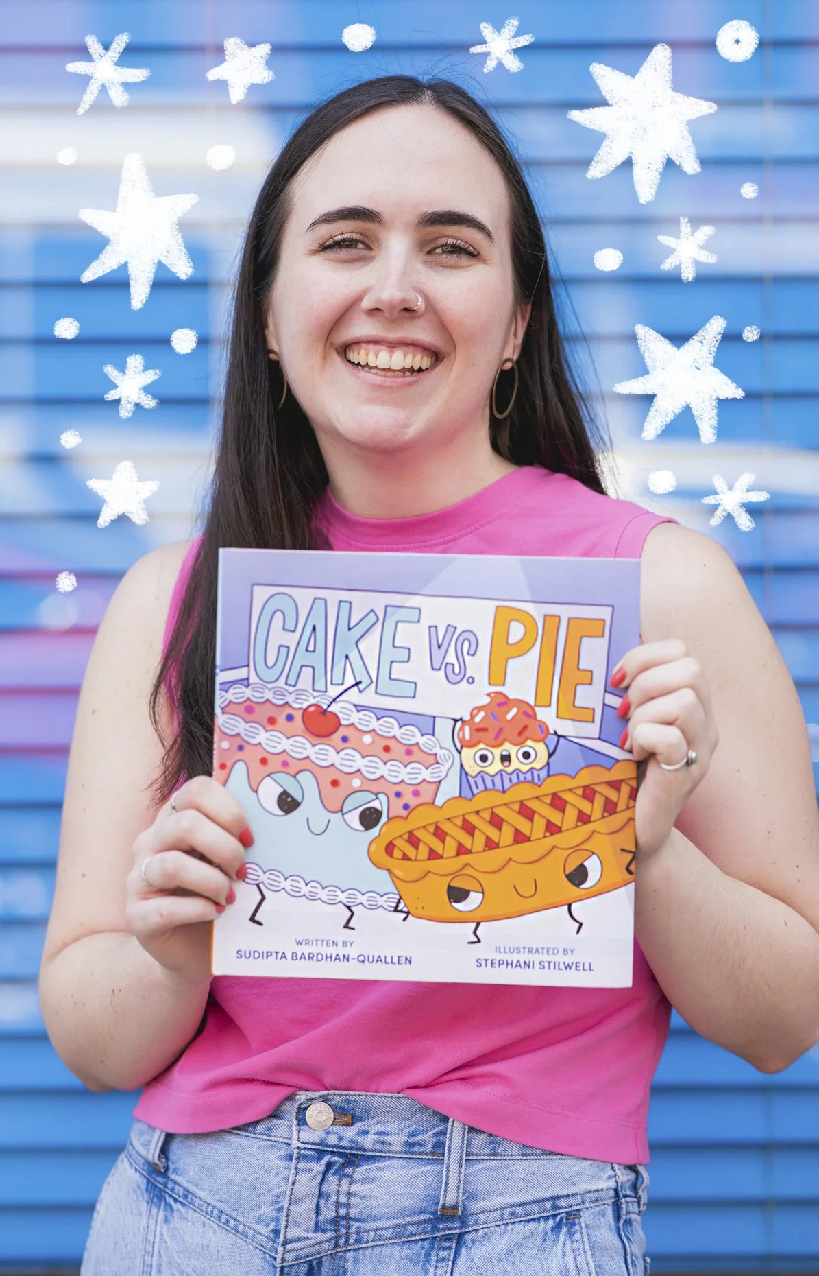 A photo of Steph smiling. She has long dark hair, is wearing a pink sleeveless top, and is holding a children's book titled 'Cake vs. Pie' in front of a blue background with drawn white stars.