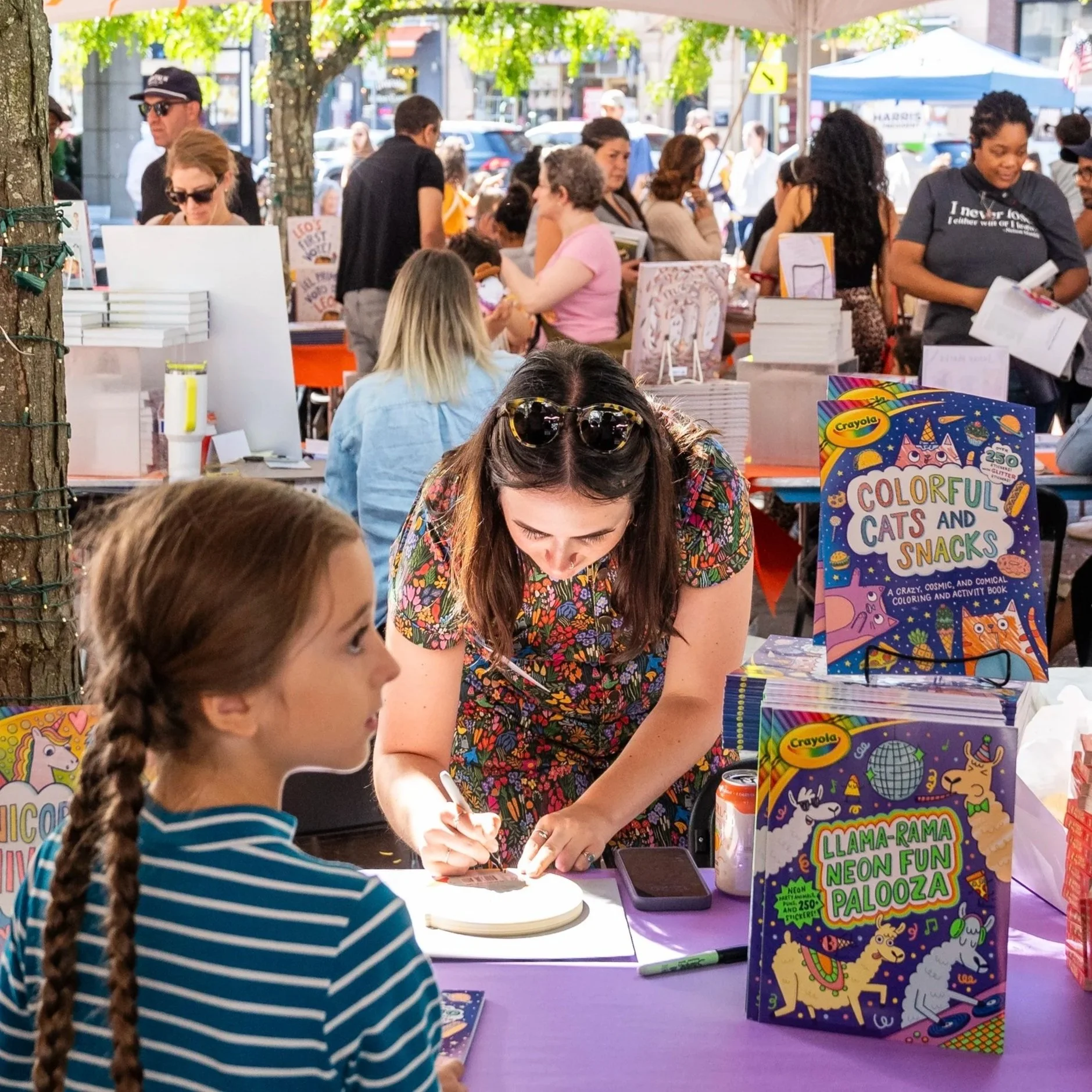A woman signing a book at a book event while a girl with braided hair and a striped shirt looks on. There are colorful books on the table, including titled 'Colorful Cats and Snacks' and 'Llama-Rama Neon Fun Palooza'. In the background, a crowd of people is at outdoor booths.