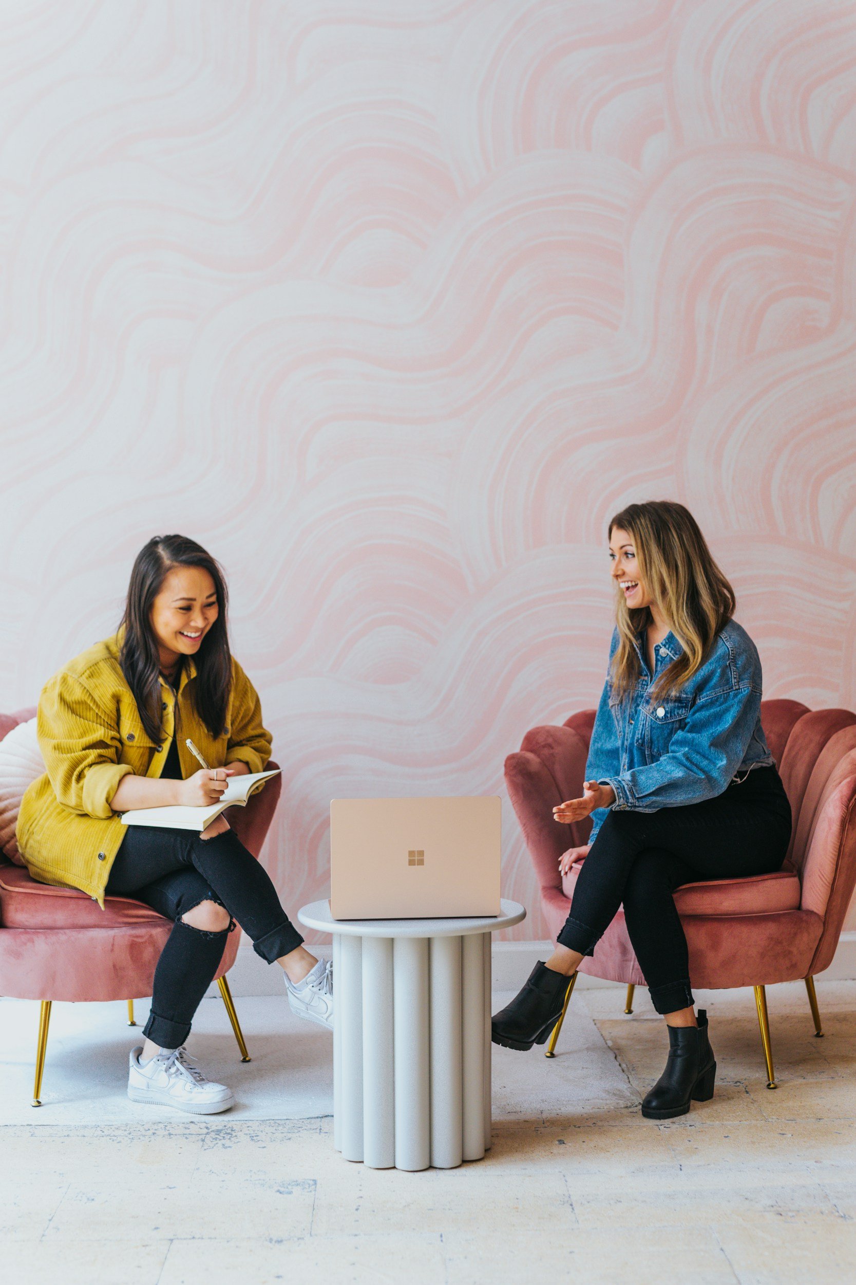 Two women sitting on pink velvet chairs having a conversation, with a white laptop on a small round table between them, in a room with pink and white abstract patterned wall.