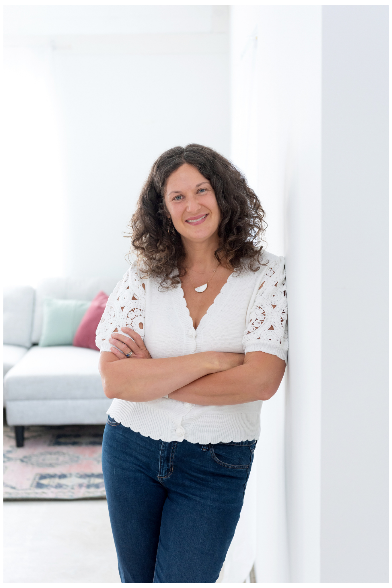 Sasha McDowell, a white woman with curly brown hair smiling and leaning against a white wall in a bright, modern living room with a sofa in the background.