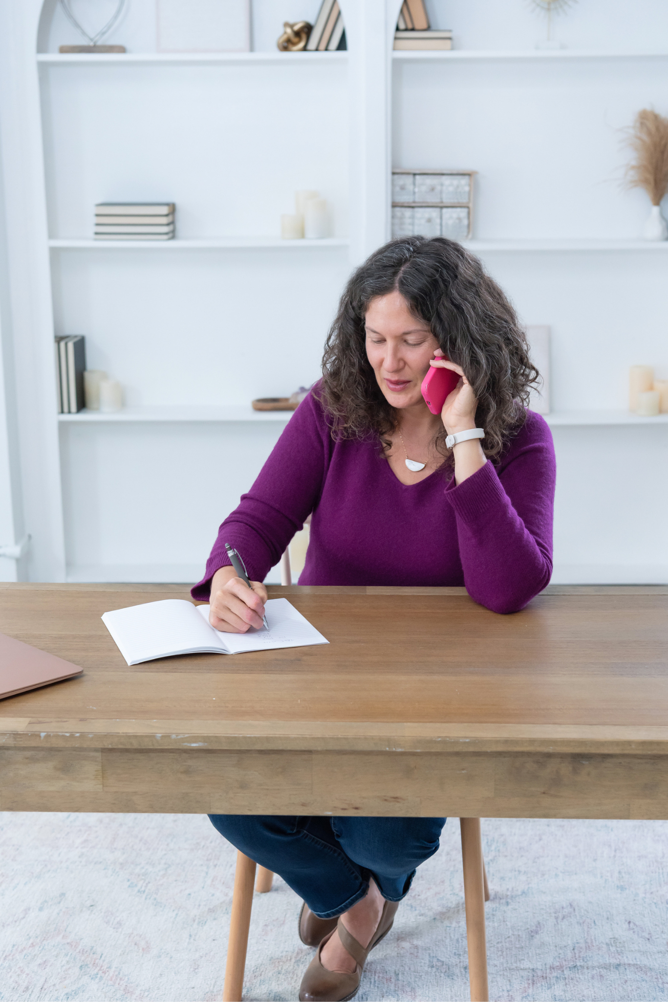 Sasha McDowell, a white woman with curly brown hair, wearing a purple sweater, is sitting at a wooden table writing in a notebook with a pen. She is talking on a pink phone.