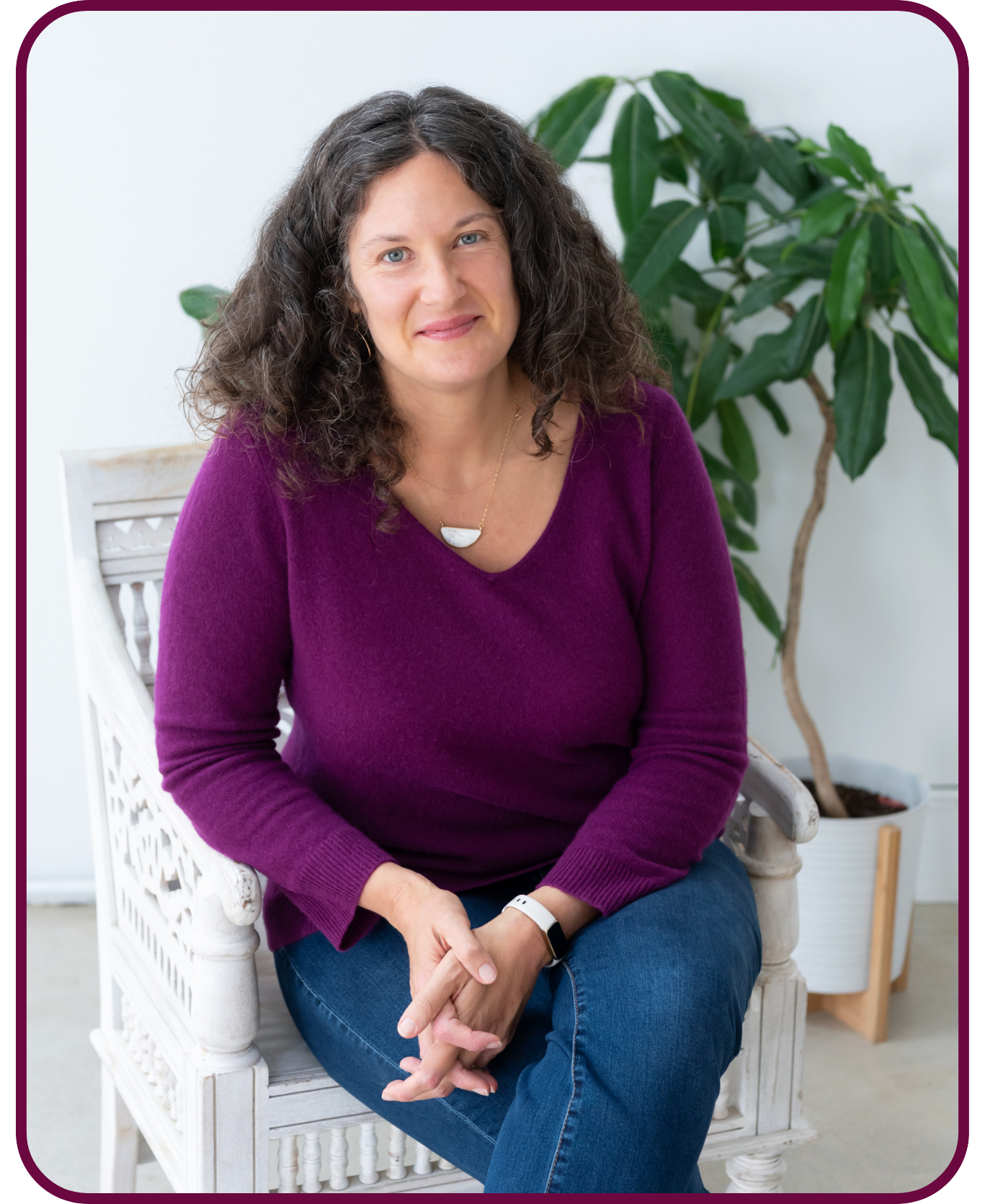 Sasha McDowell, a white woman with curly brown hair and blue eyes sitting on a white wooden chair, wearing a purple sweater and blue jeans, with a large green plant in the background.