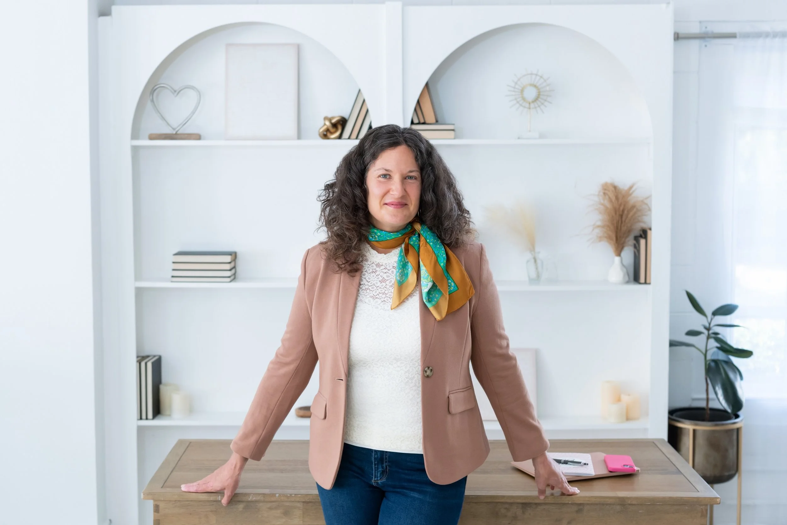 Sasha McDowell, a white woman wearing a tan blazer, green and orange scarf, white top and jeans leans against a desk with white bookshelves behind her.