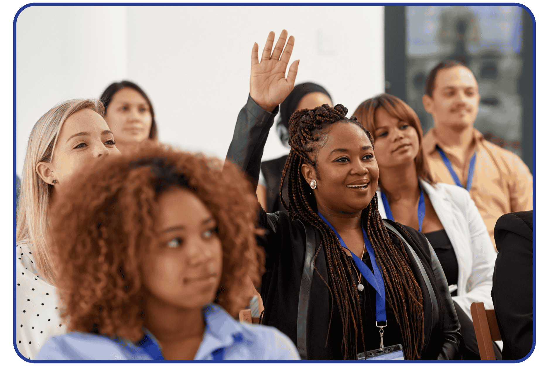 A diverse group of women at a conference, with one woman raising her hand to ask a question or participate, wearing a lanyard and smiling.