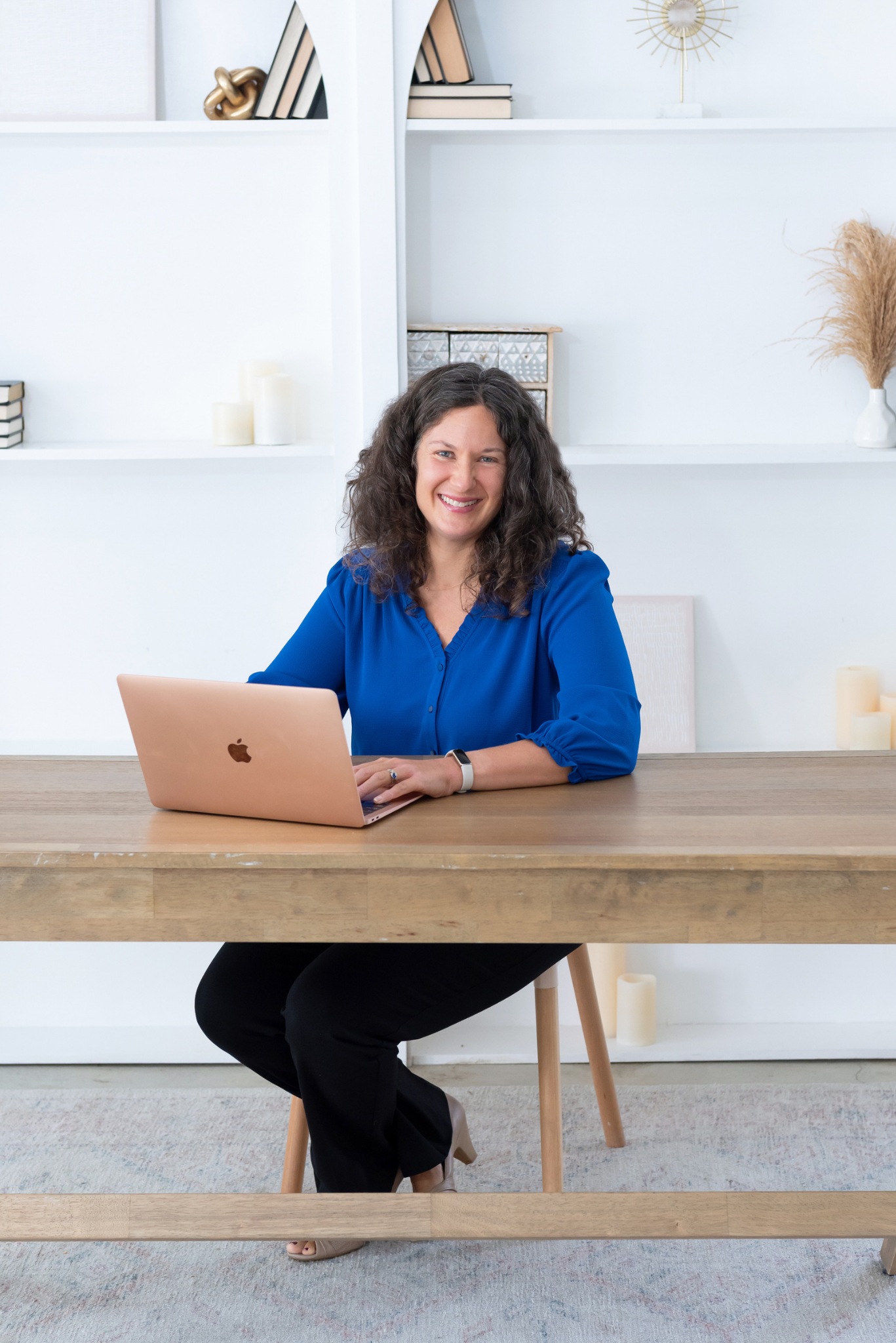 Sasha McDowell, a white woman with curly hair and bright blue bouse smiling and sitting at a desk with a laptop, in a modern bright home office with white shelves and decorative items.