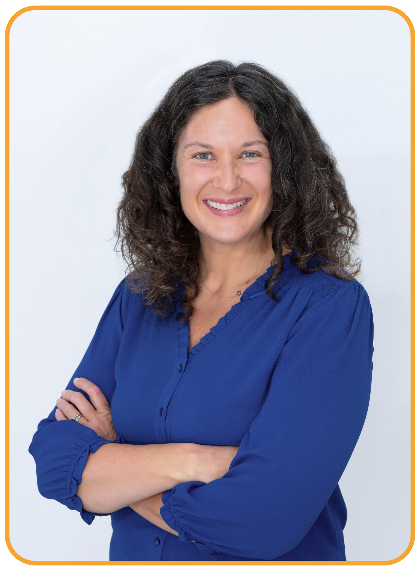 Sasha McDowell, a white woman with curly dark hair, wearing a blue blouse with rolled-up sleeves, smiling, with arms crossed, standing against a white background.