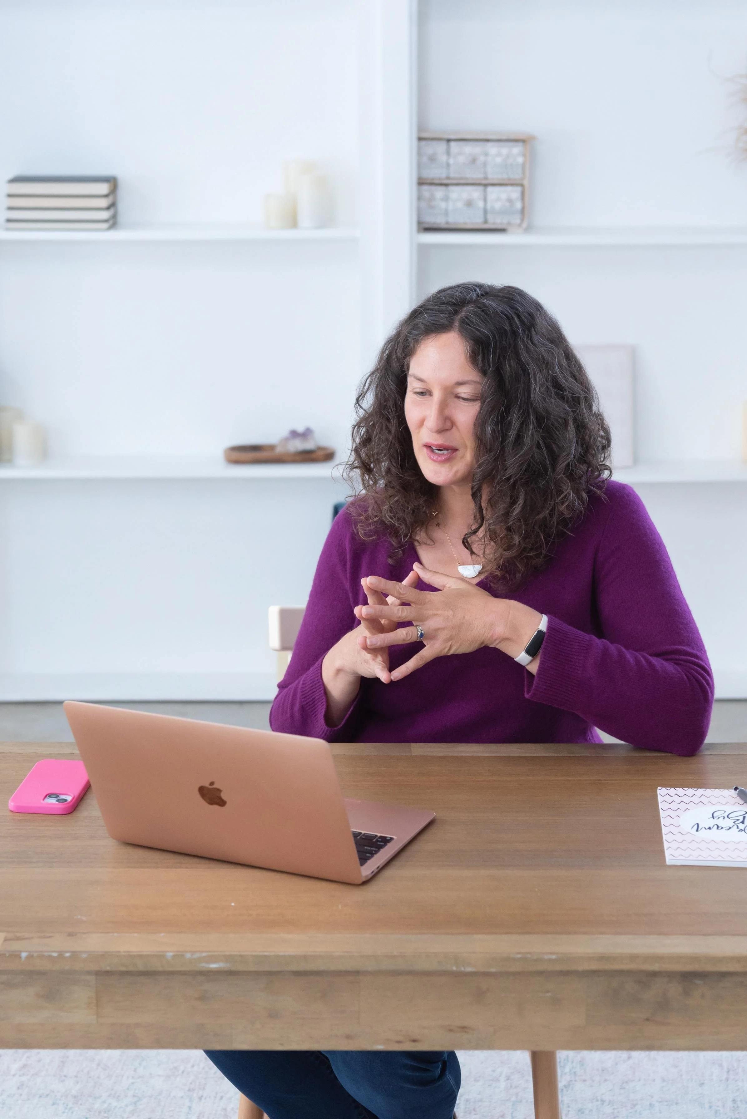 Sasha McDowell, a white woman with curly dark hair wearing a purple blouse sitting at a desk and gesturing at a blush laptop on a video call.  Next to her is a bright pink phone, she's in a bright room with white walls and shelves.