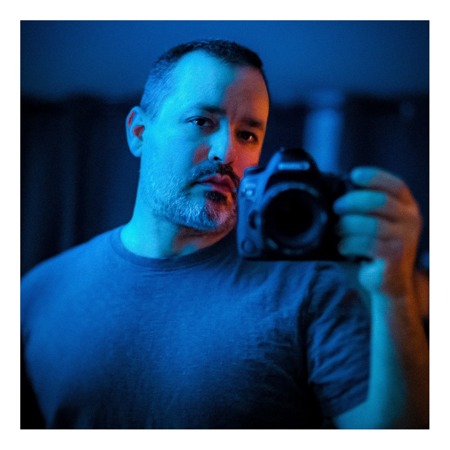 Self Portrait selfie with a camera, illuminated with blue light, in a dimly lit room.