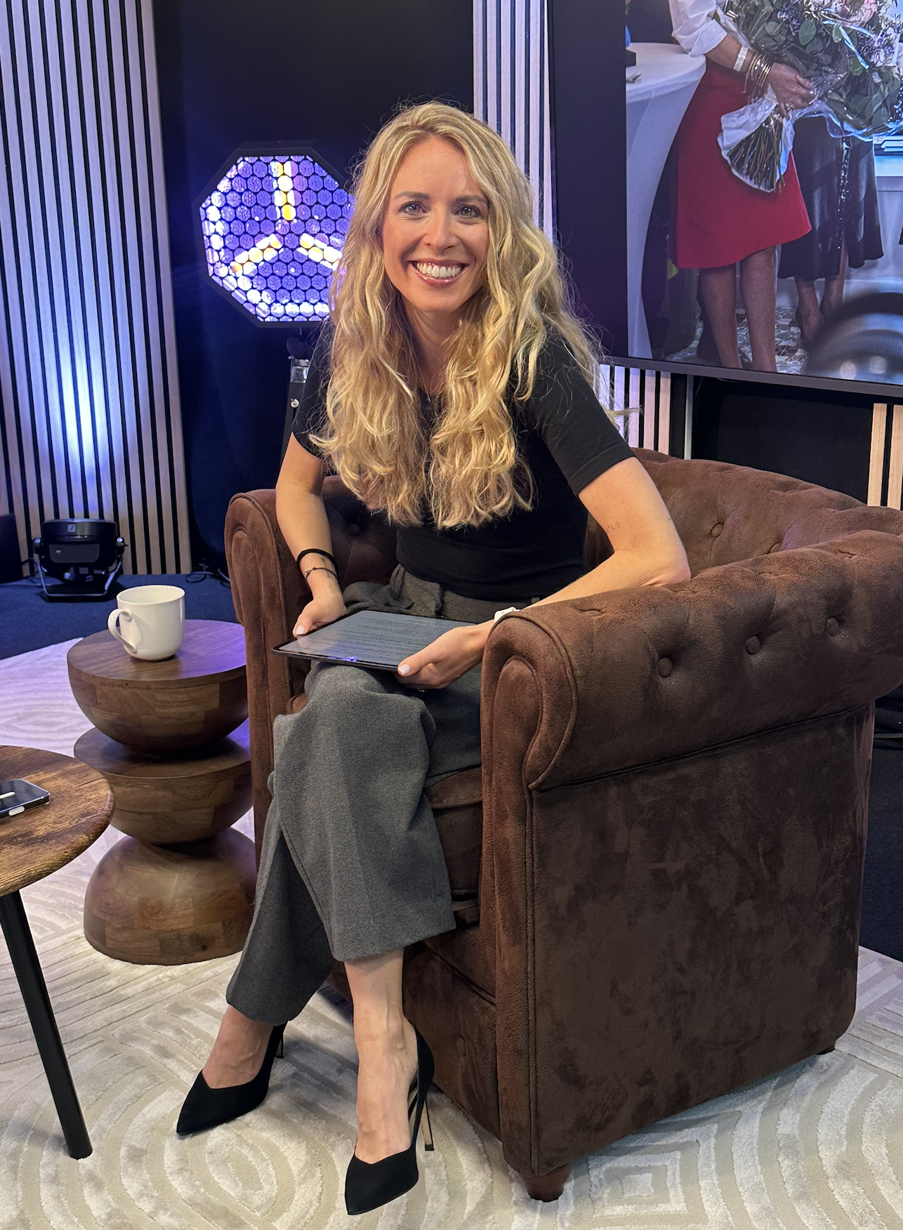Une femme aux cheveux bouclés blonds souriante, assise sur un fauteuil en cuir marron, tenant une tablette dans les mains. Elle porte une robe noire, des pantalons gris et des talons noirs. À côté d'elle, une tasse blanche est posée sur une petite table en bois. Dans le fond, un écran affiche une image floue de personnes.