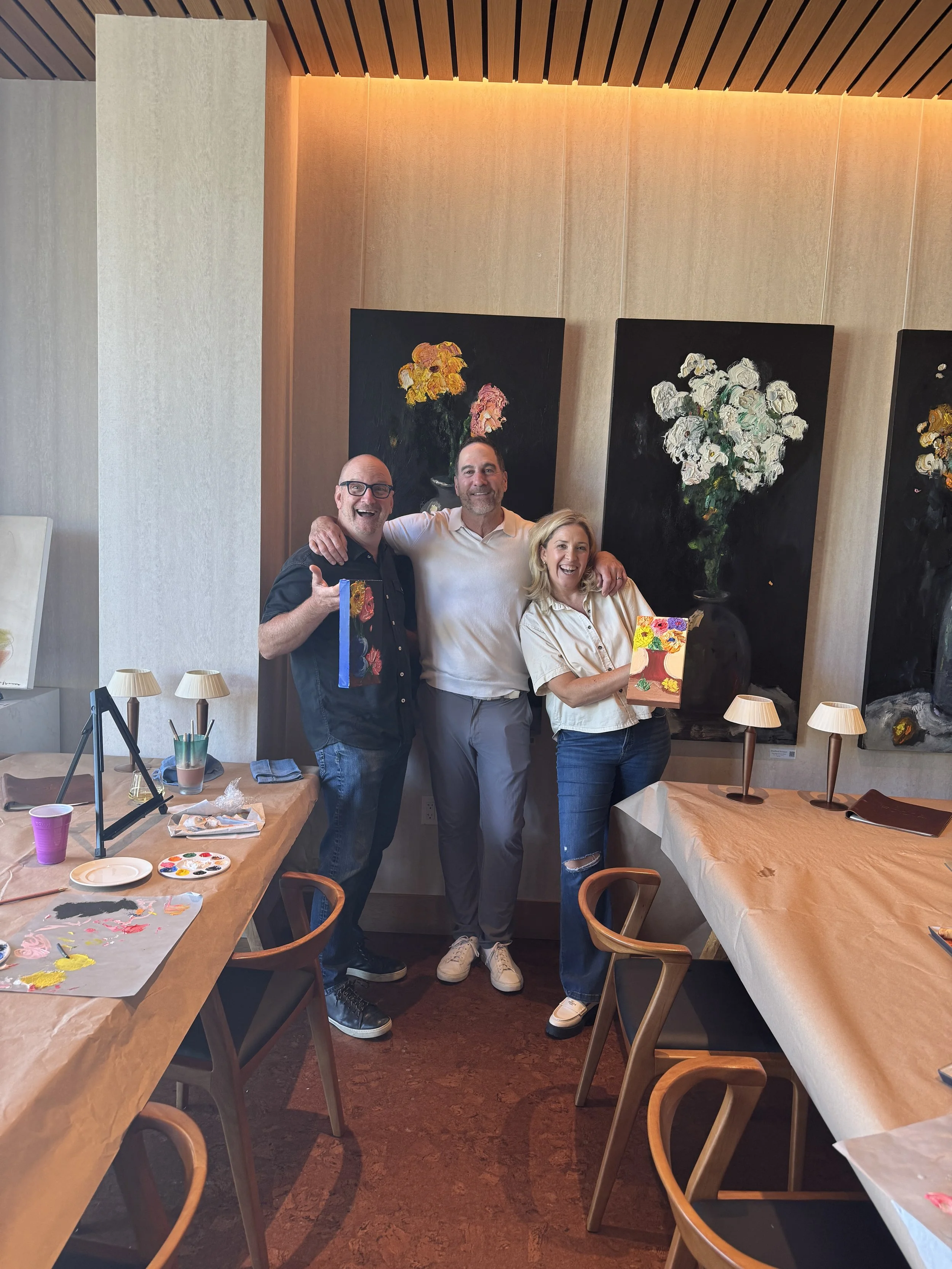 Three guests in aprons holding their completed colorful floral paintings, standing in front of large glass doors with art supplies on the table in the foreground.