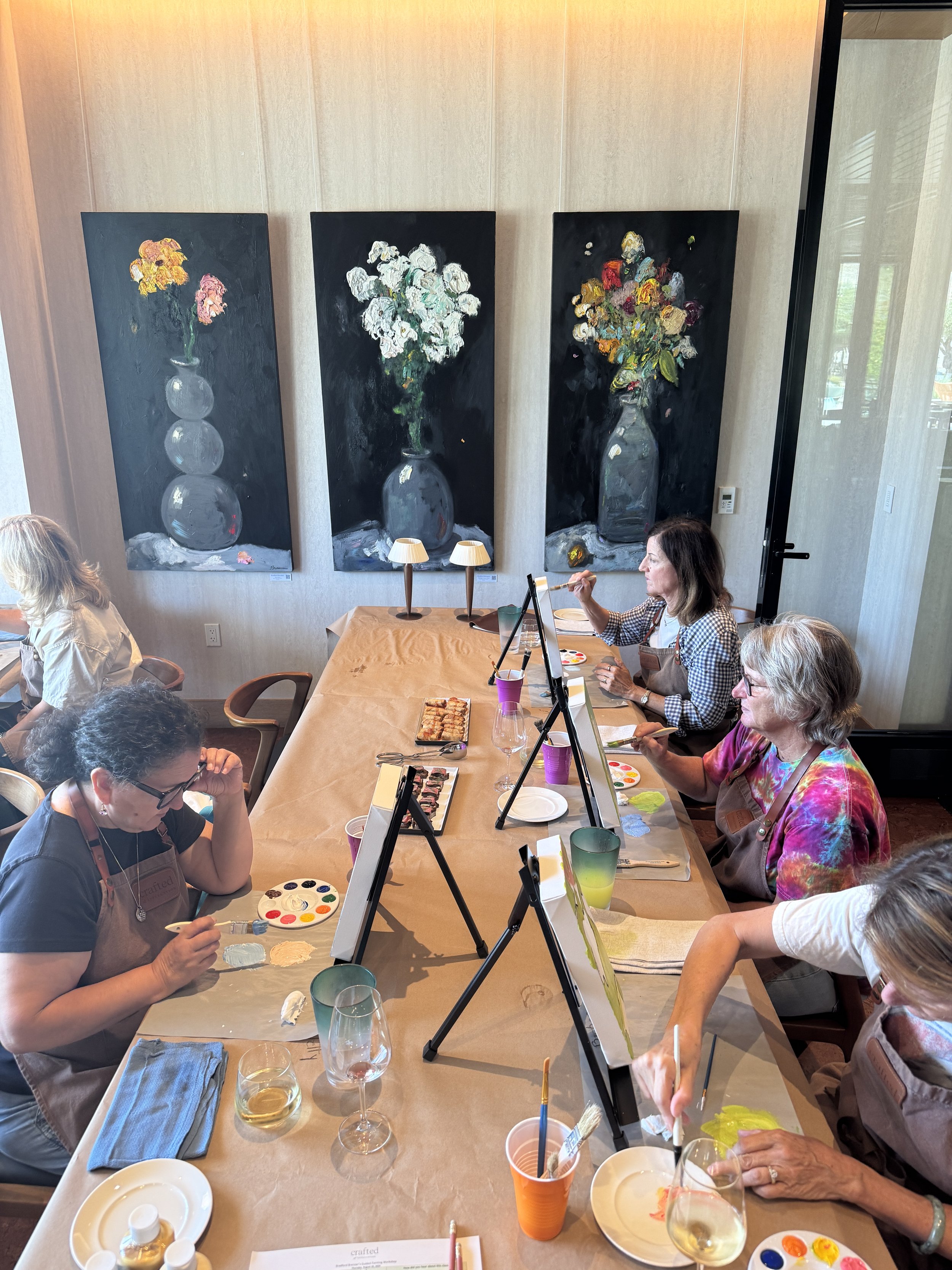 Small group of guests painting floral canvases at a table while an instructor assists, with food, drinks, and large floral artwork in the background.
