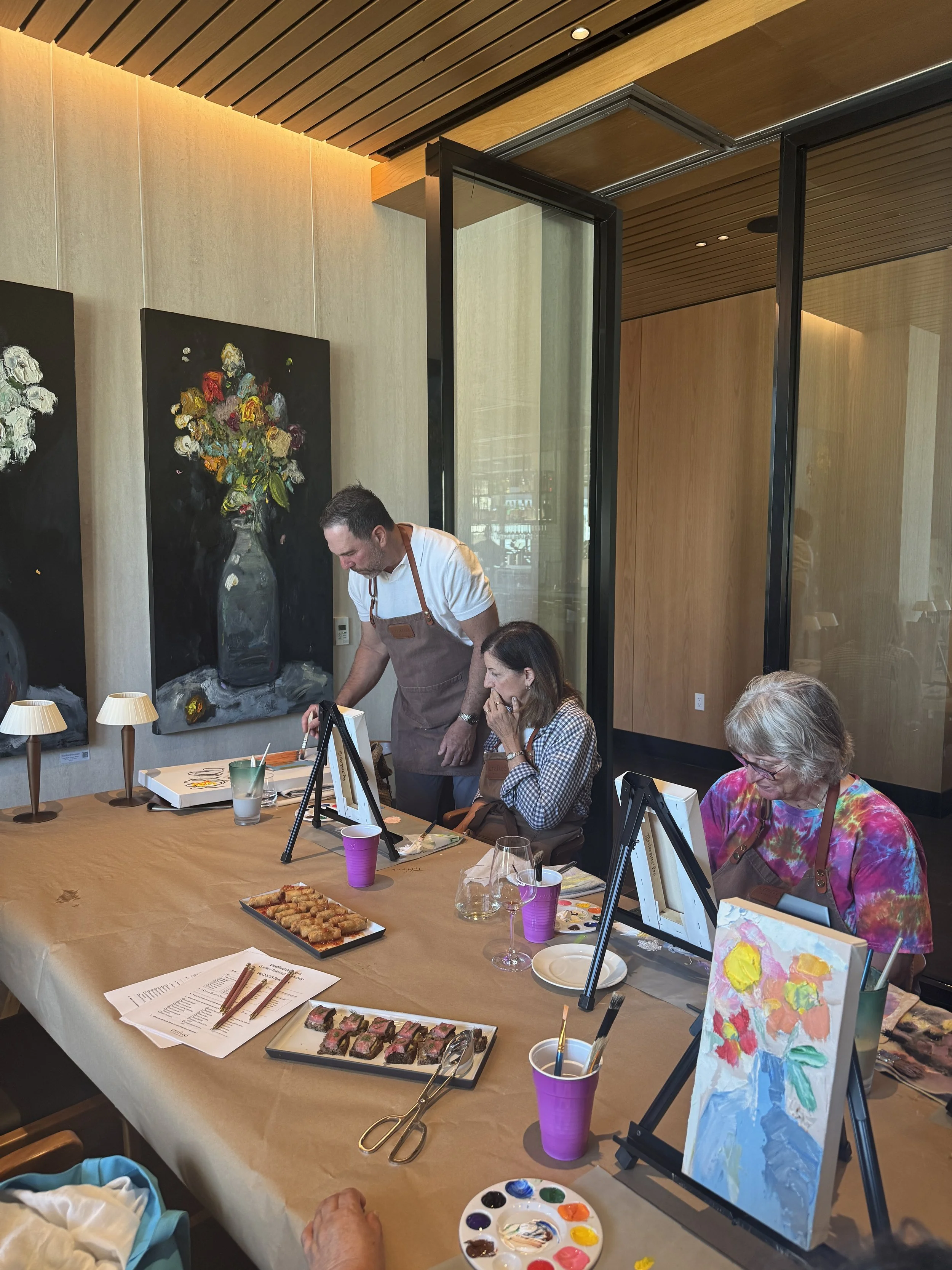Guests seated at a long table working on small floral canvases during the paint‑with‑the‑artist workshop, with large flower paintings displayed on the wall behind them.