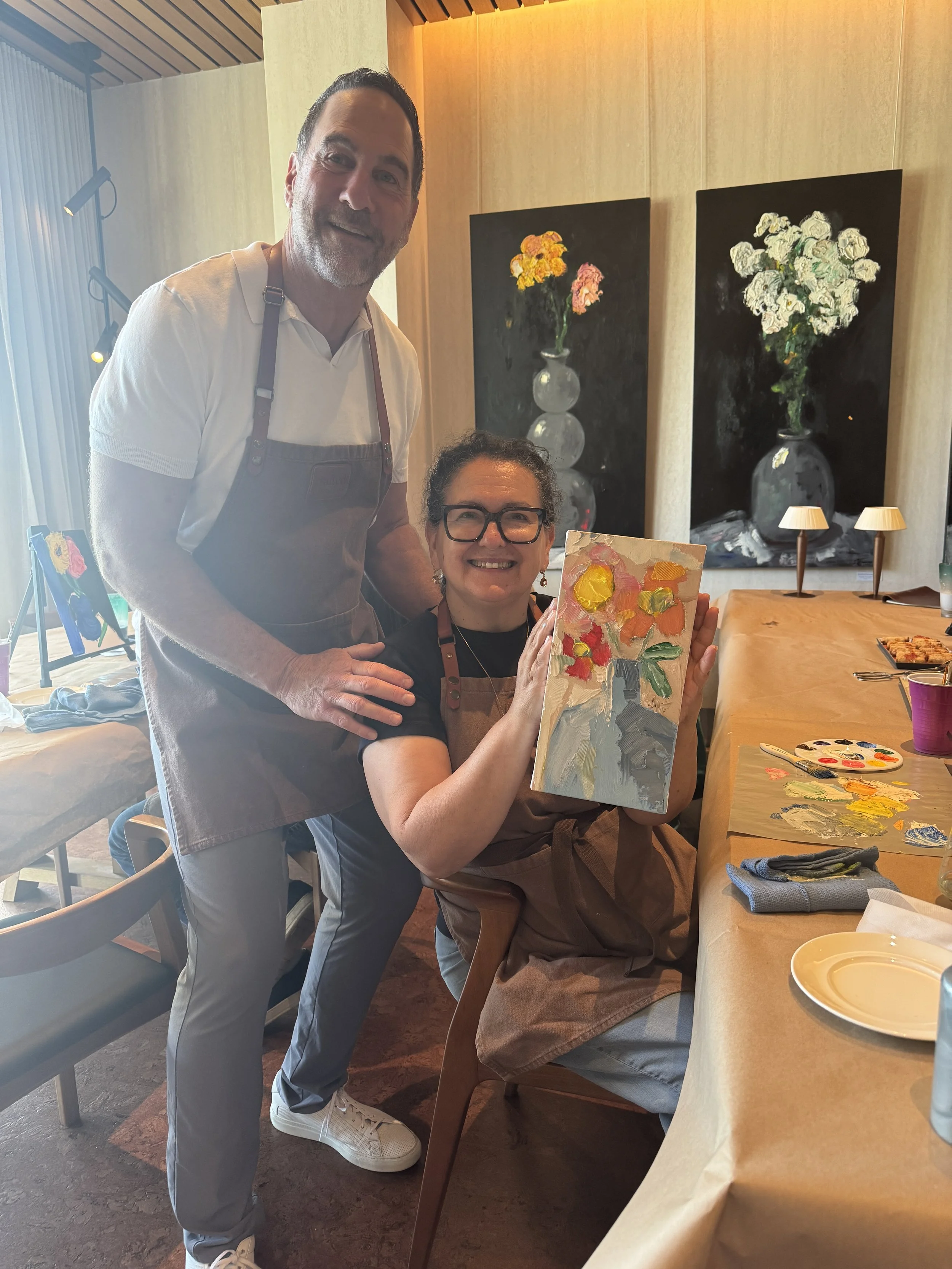 Two participants in aprons smiling as one holds up a finished floral painting, surrounded by art supplies and large flower artworks on the wall.