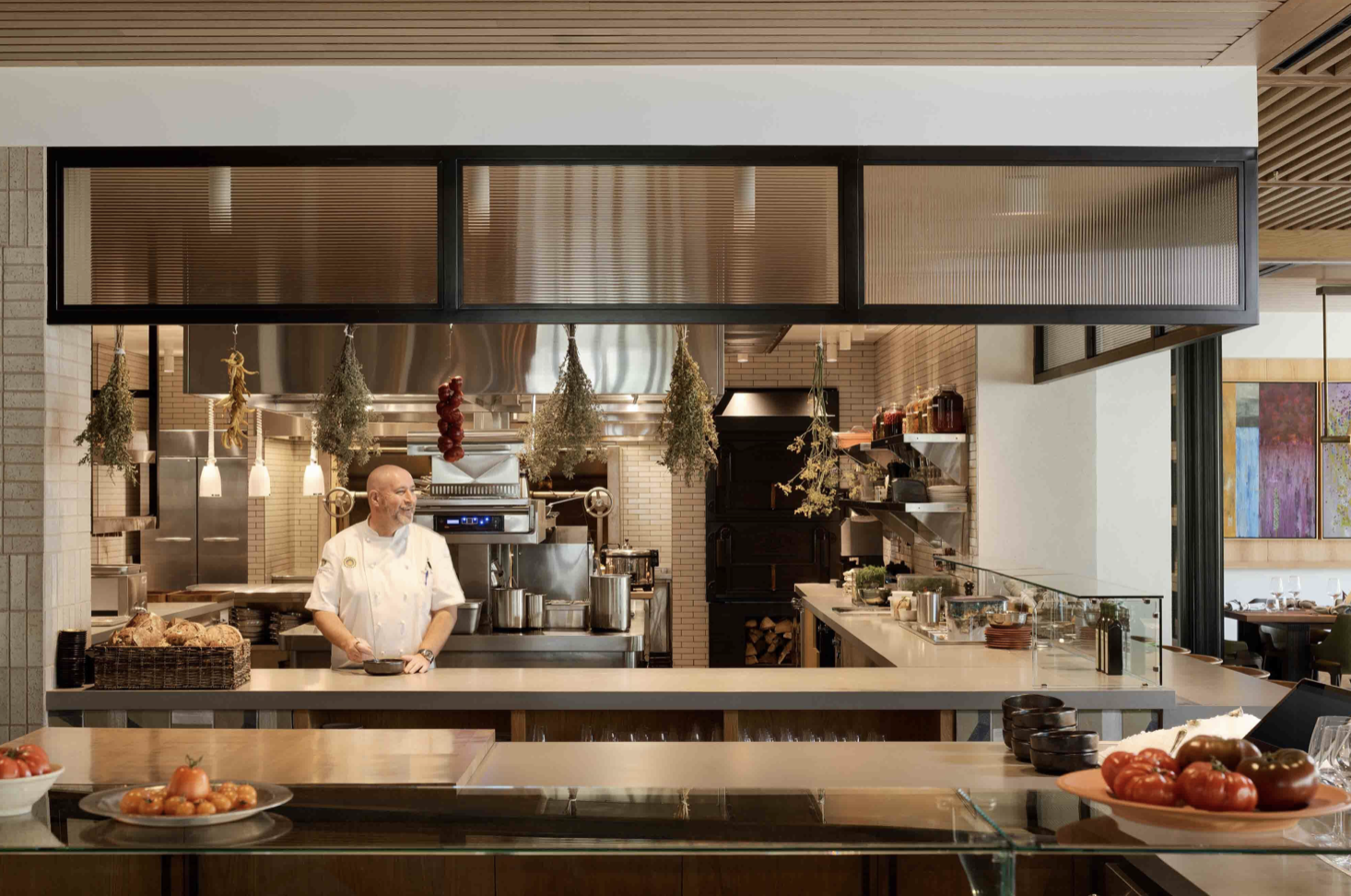 Professional chef in a modern open kitchen preparing food, surrounded by fresh ingredients, bread, and stainless-steel appliances.