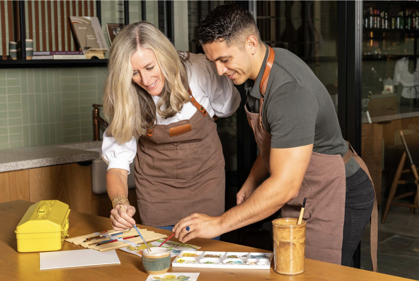 Two people wearing aprons working on watercolor paintings at a wooden table with brushes, paints, and art supplies.
