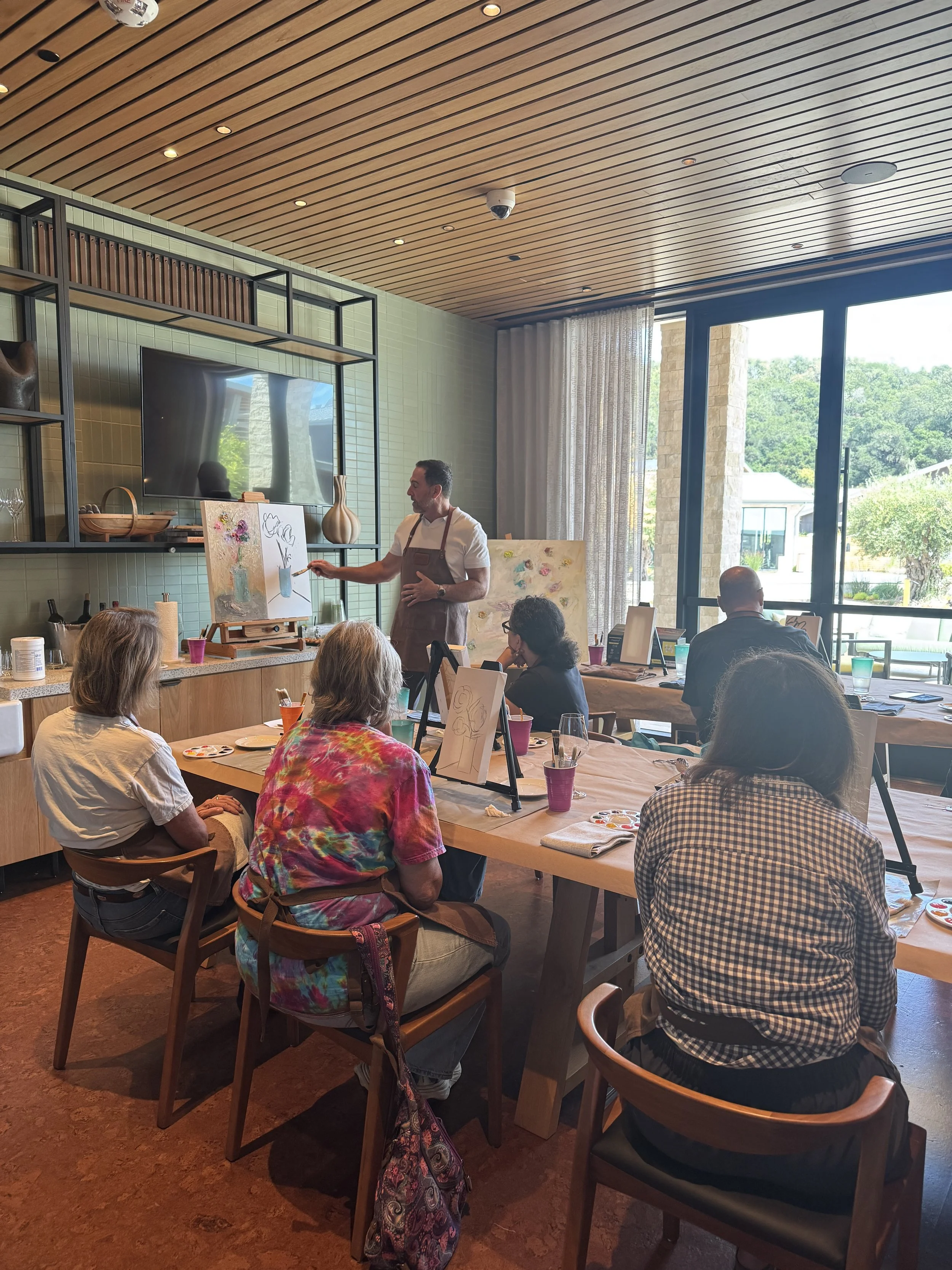 Guests seated at a long table during the paint‑with‑the‑artist workshop, following a demonstration as the instructor paints on an easel in a bright, modern room.