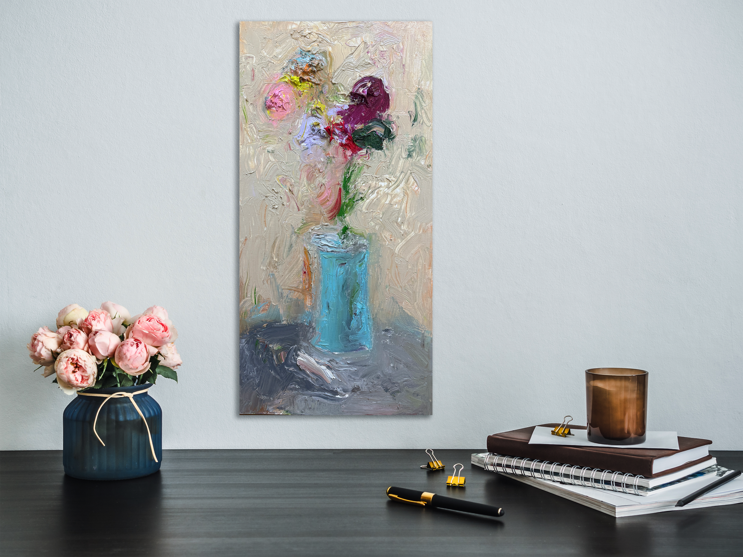 Modern interior with a floral still‑life painting hanging above a dark tabletop styled with a blue vase of pink roses, stacked books, and a candle, creating a warm, art focused vignette.