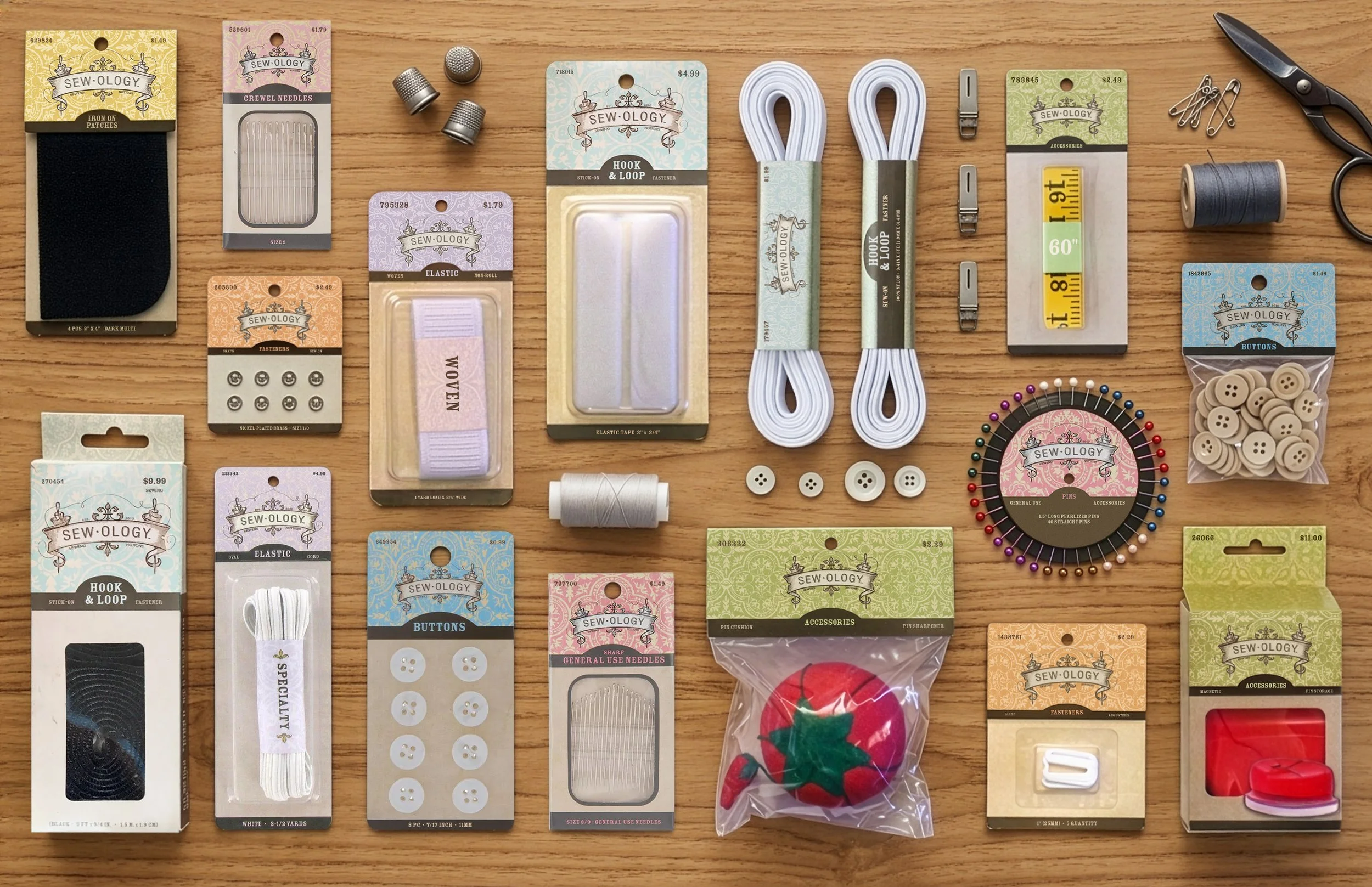 Sewology sewing notions flat lay featuring sewing needles, buttons, elastic cord, hook-and-loop fasteners, thread, pins, safety pins, measuring tape, thimbles, scissors, pincushion, and iron-on patch materials on a wood surface.