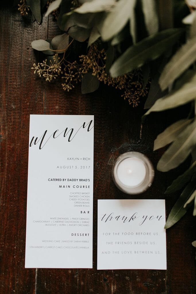 Printed wedding menu and thank-you cards styled with greenery and candle on a wooden reception table