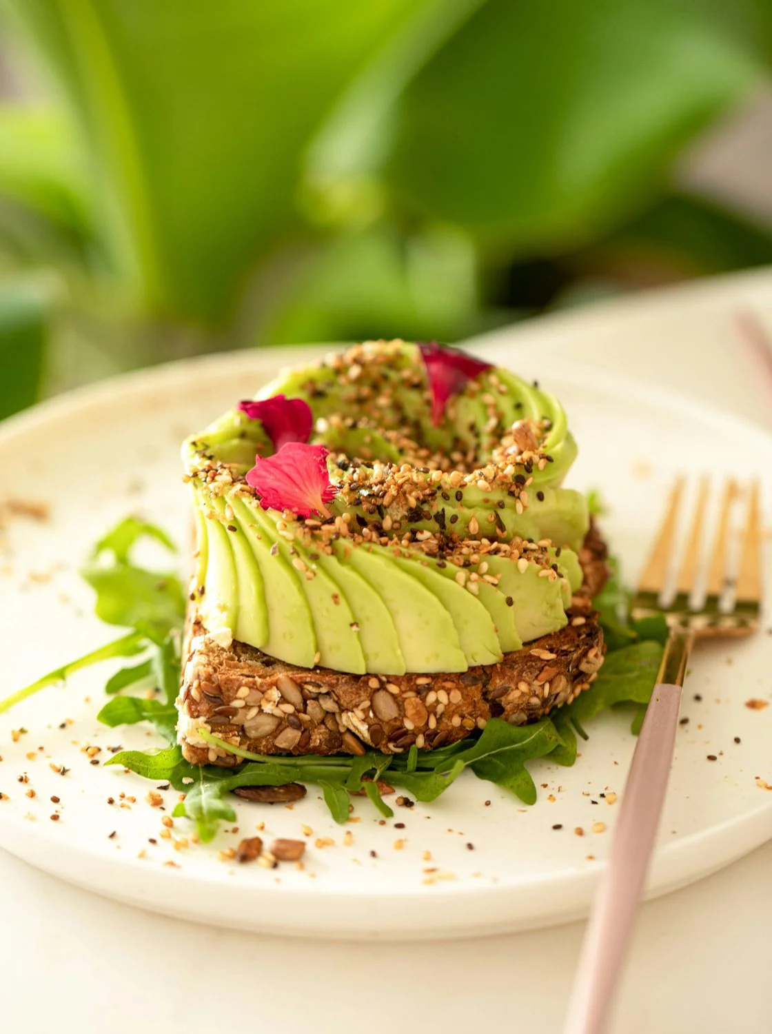 beautiful editorial shot of a piece of whole grain toast with a ripe avocado fanned out across it, garnished with several small, bright tropical flowers.