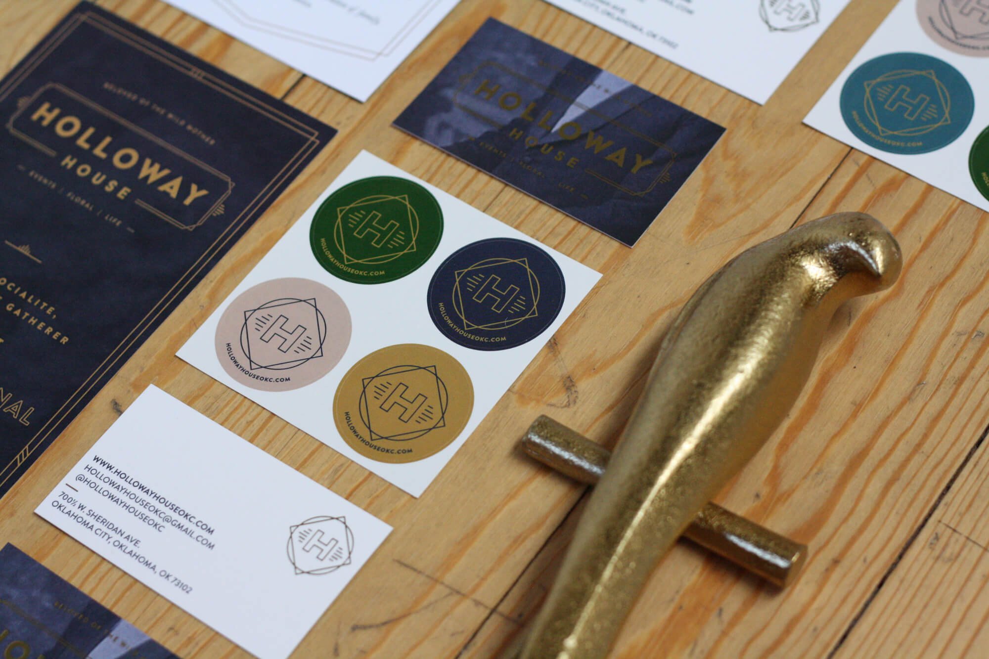 Holloway House branded marketing materials displayed on a wooden tabletop, including business cards, logo stickers, brochures, and promotional stationery alongside a gold decorative parrot.