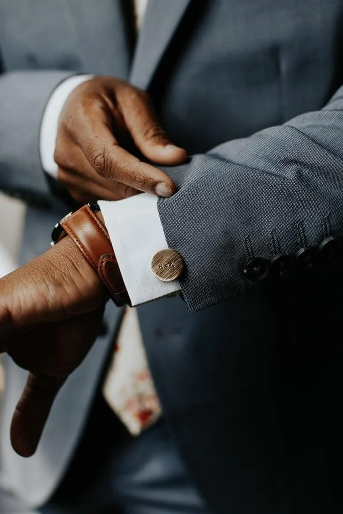 Close-up of a man adjusting a cufflink on a gray suit jacket, wearing a brown leather watch