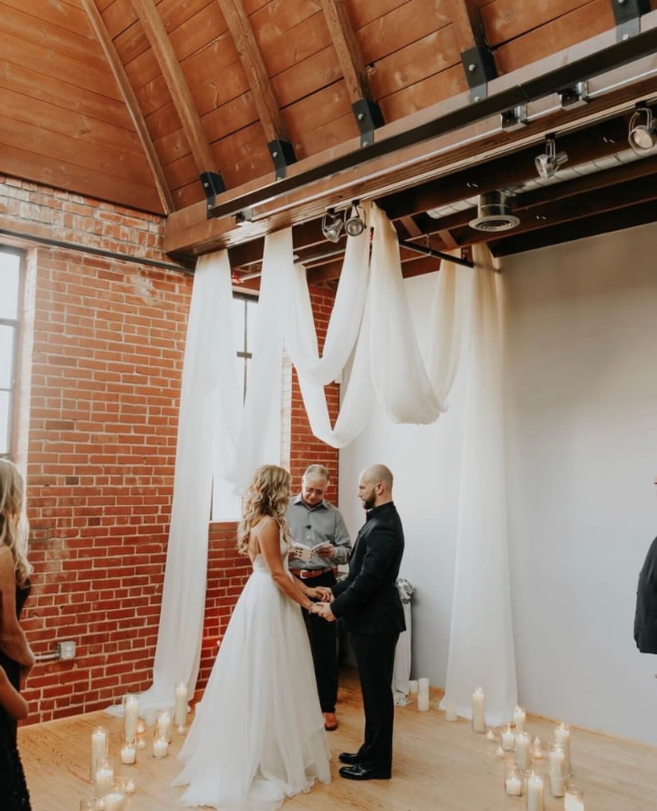 Event venue interior showing wood beam ceiling and exposed brick with draped fabric ceremony installation and candlelight