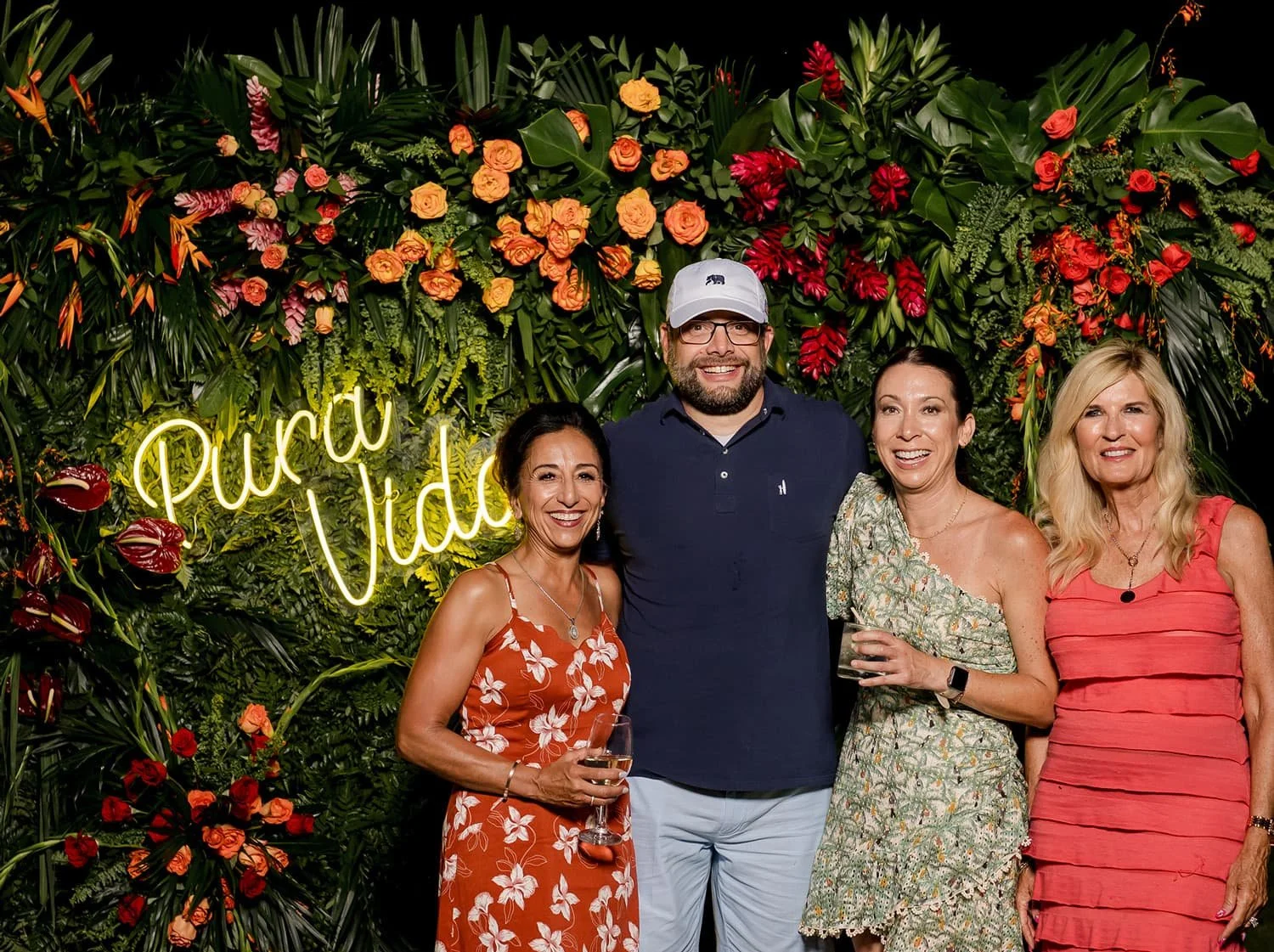 Lifestyle event photography capturing attendees posing in front of a lush tropical floral backdrop with a glowing “Pura Vida” neon sign, showcasing an immersive brand experience with vibrant greenery and colorful flowers.