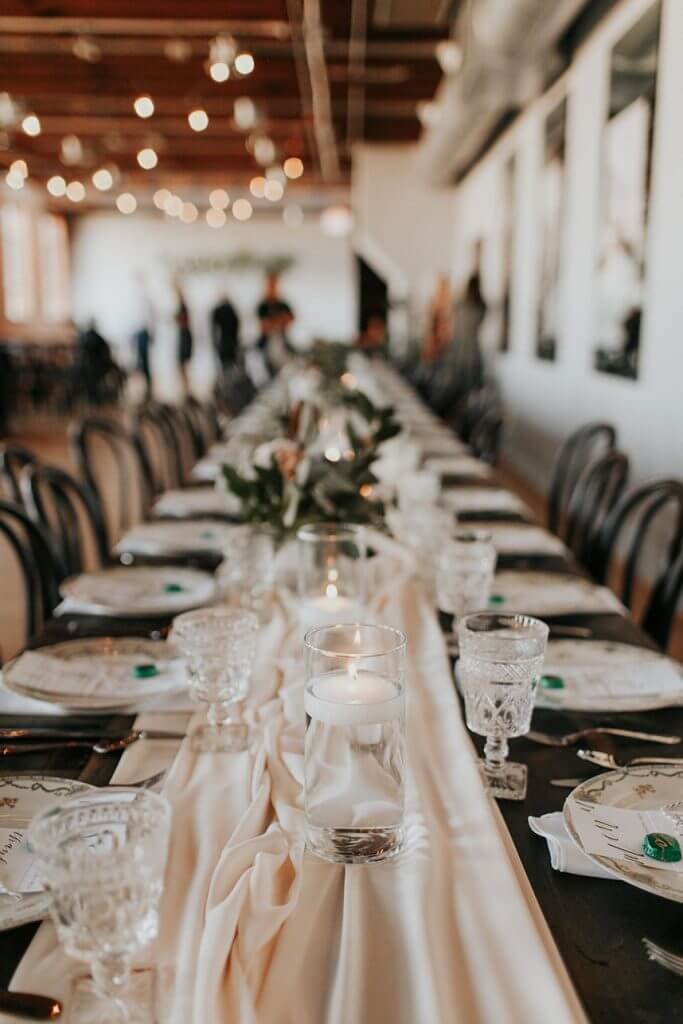 Event venue interior with long reception table, candlelight, and greenery under wood beams and large windows