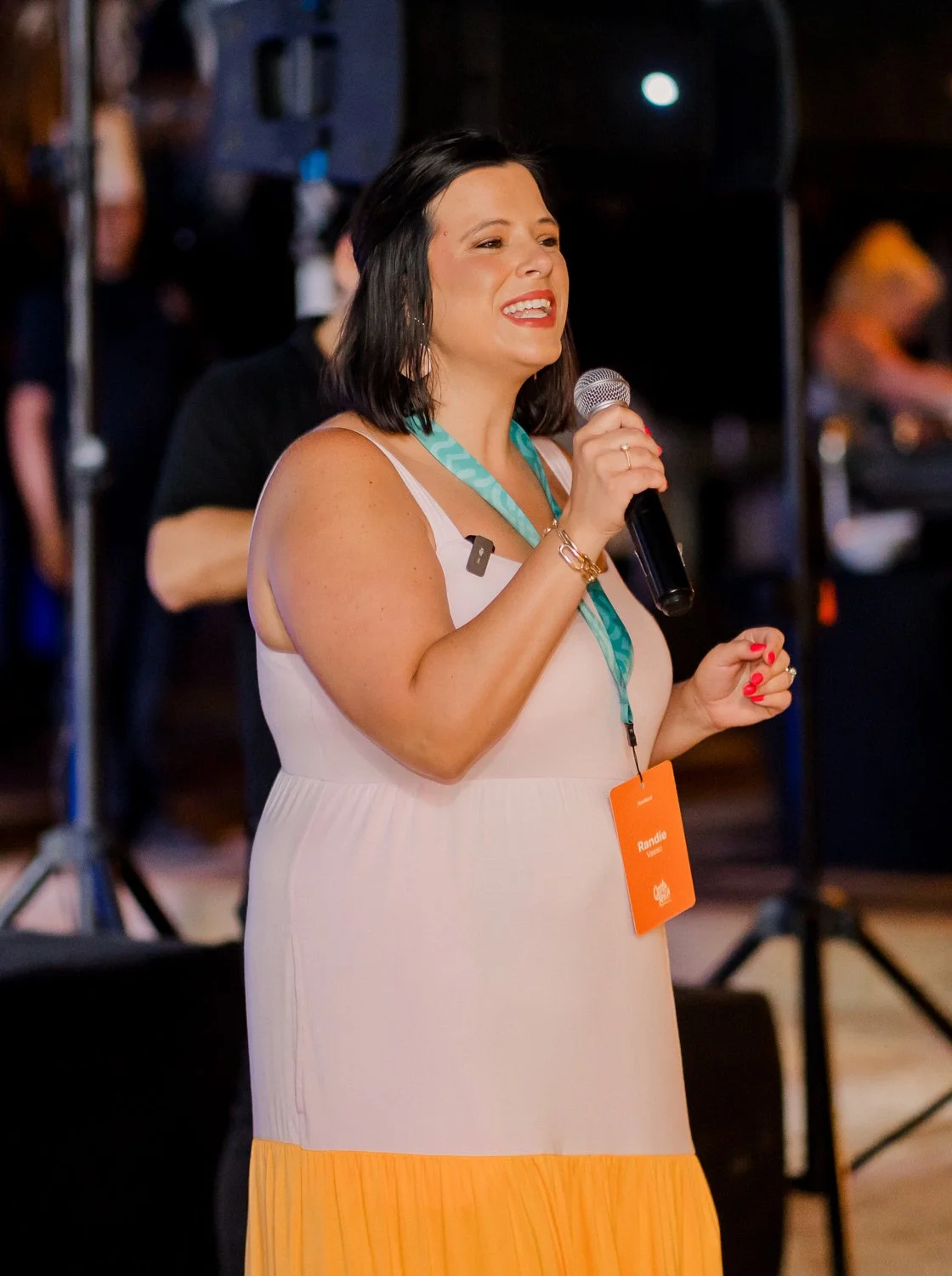 Candid event photo capturing a speaker holding a microphone during a live presentation, wearing a branded lanyard badge, with stage lighting and production setup visible in the background.